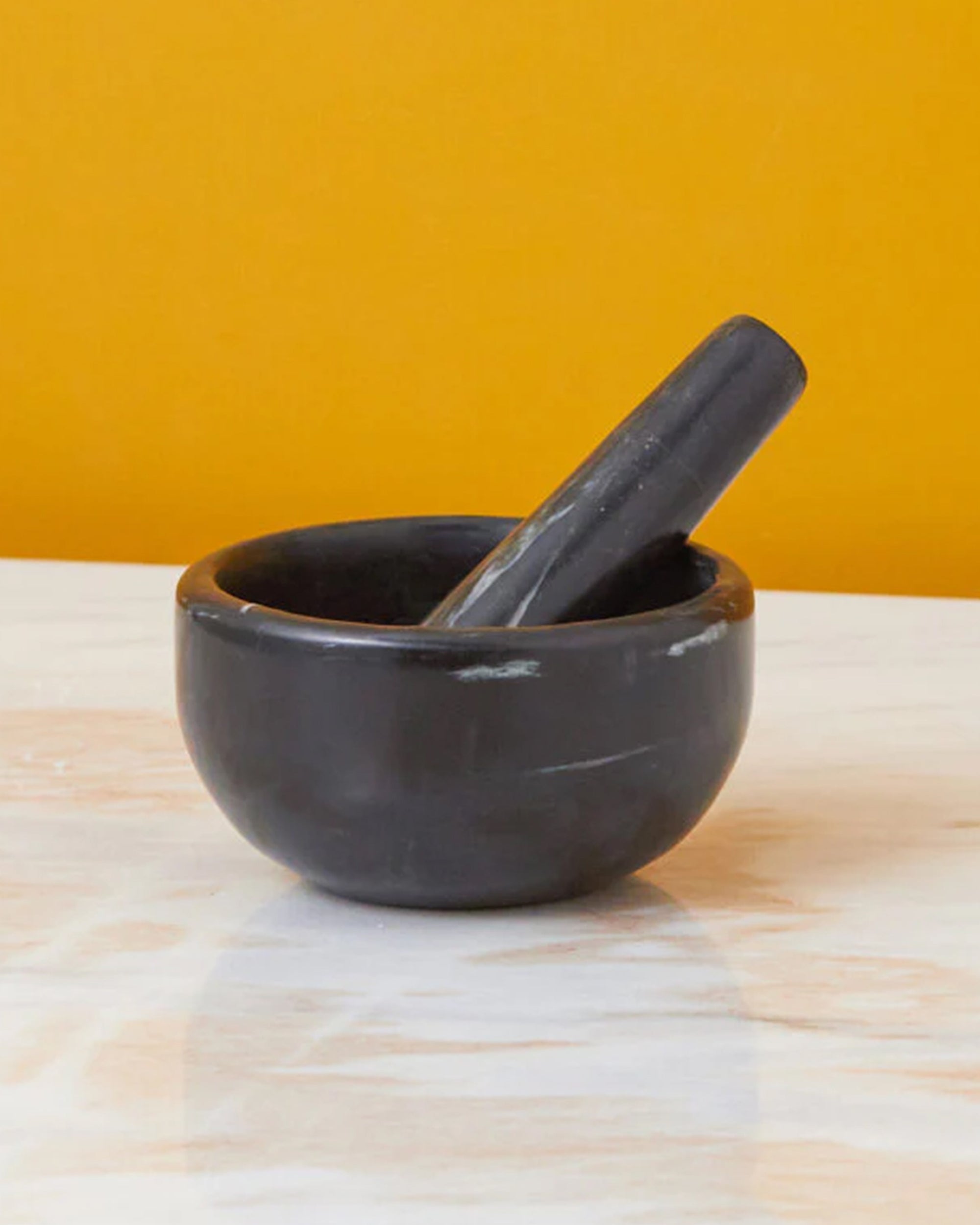 The Be Home Salerno Black Marble Mortar & Pestle, with natural striations, rests on a white marble surface against a vivid yellow backdrop.