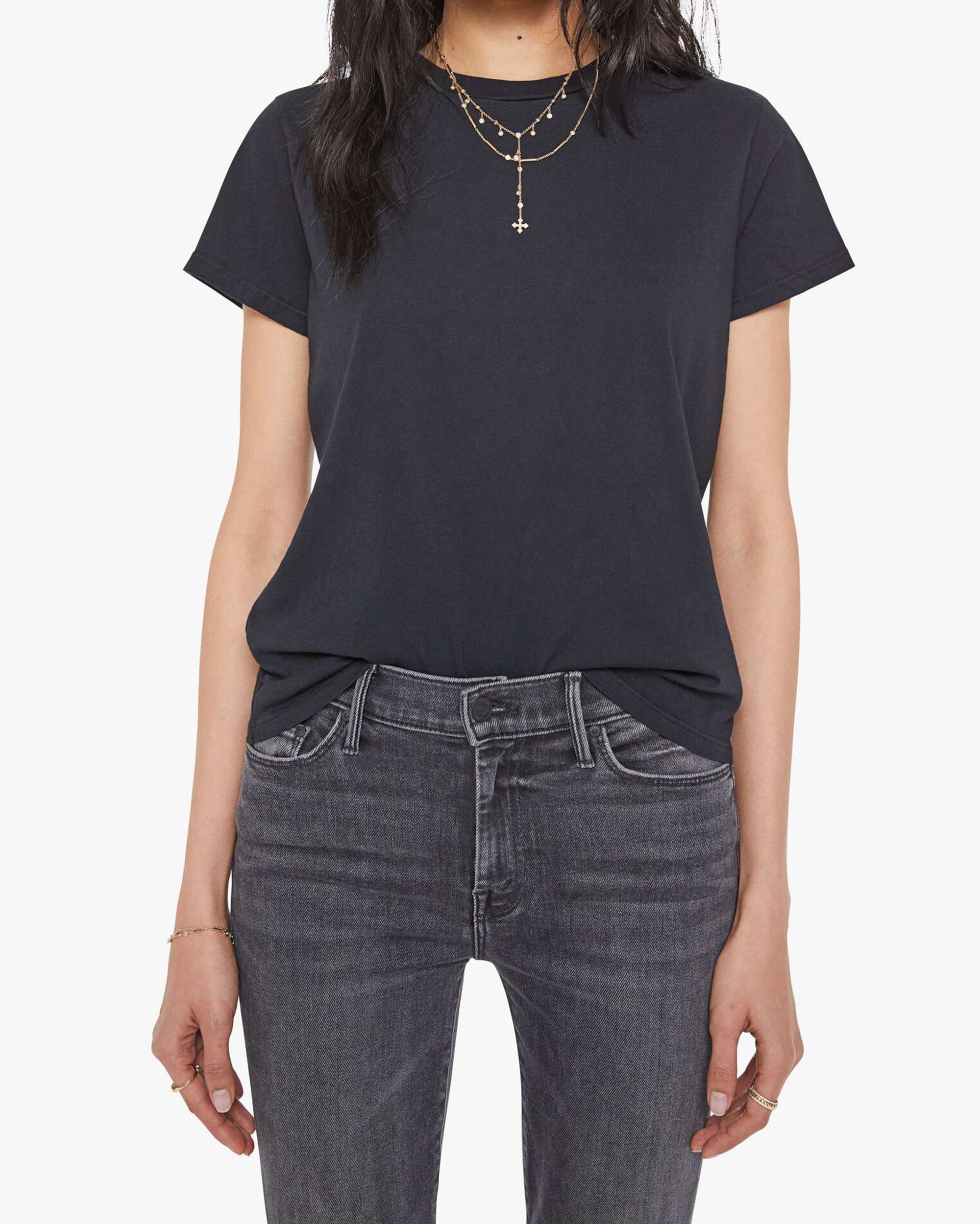 A woman wears Mother’s The Lil Goodie Goodie Tee in plain black, tucked into faded black jeans with layered gold necklaces and bracelets, standing against a white background.
