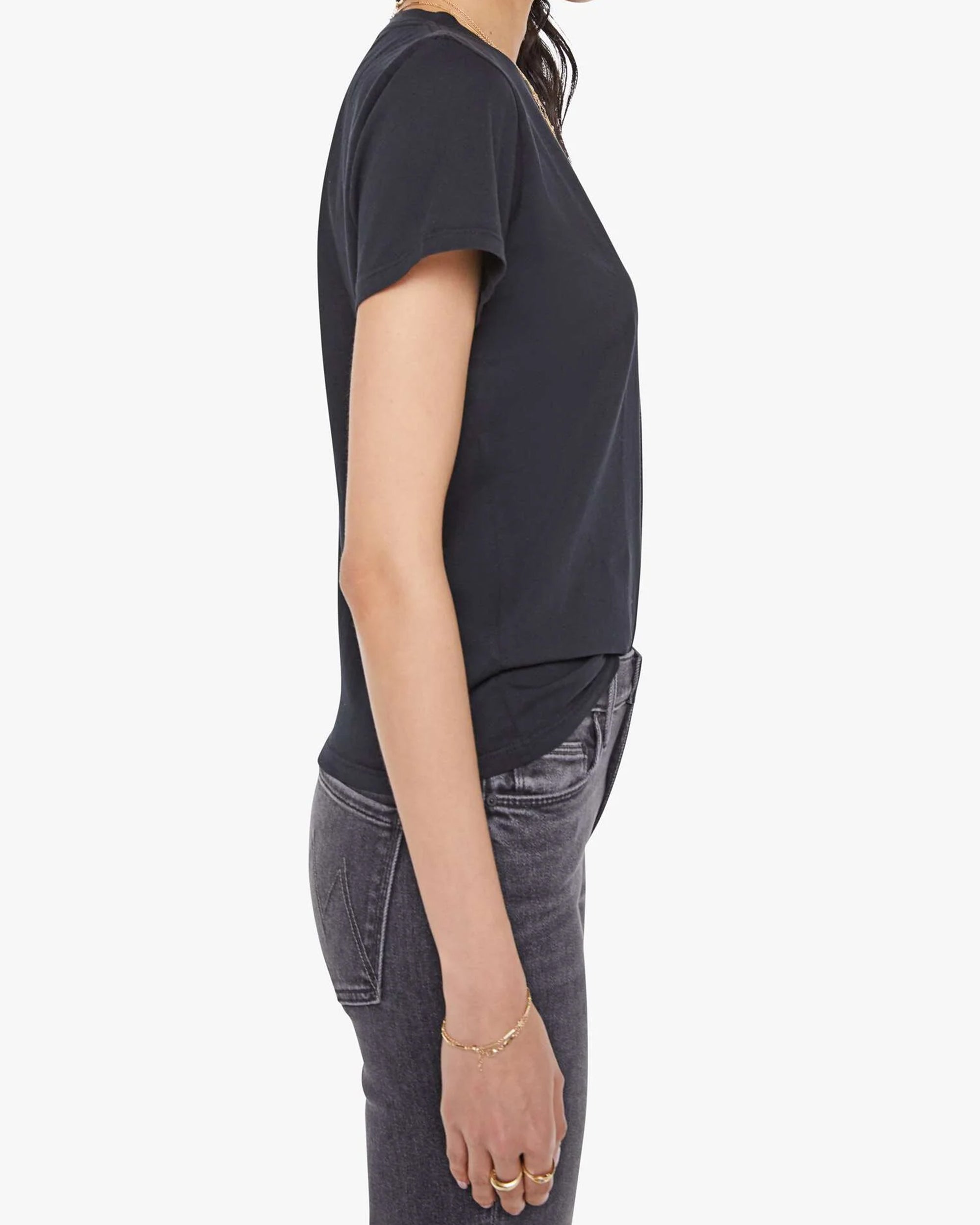 Side view of a person wearing Mother’s The Lil Goodie Goodie Tee in super soft fabric and dark gray jeans, left arm relaxed by their side, standing against a plain light background.