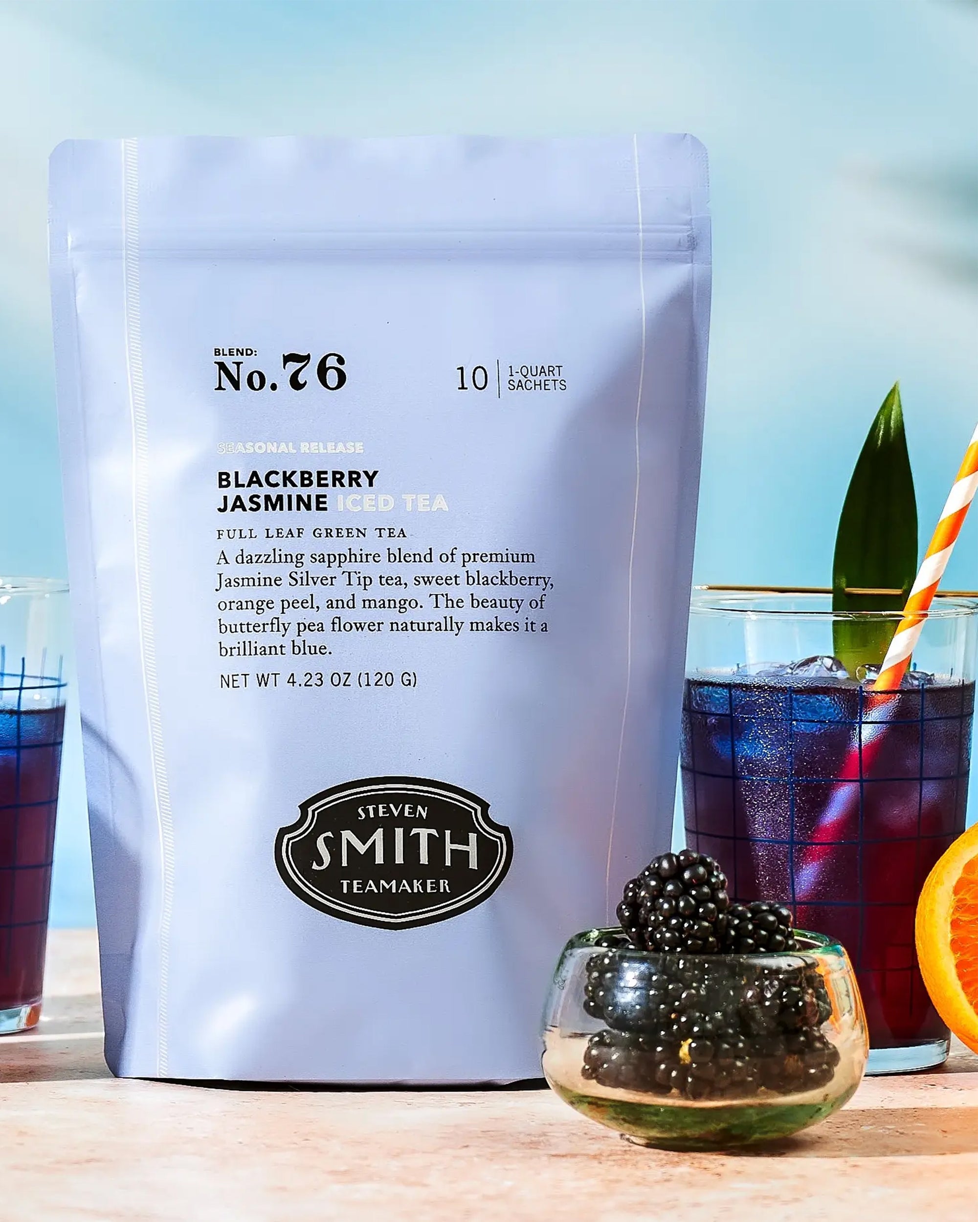 A bag of Faire Blackberry Jasmine Iced Tea stands next to a glass of iced tea with a striped straw, fresh blackberries, and an orange slice. A small bowl of blackberries is in the foreground.