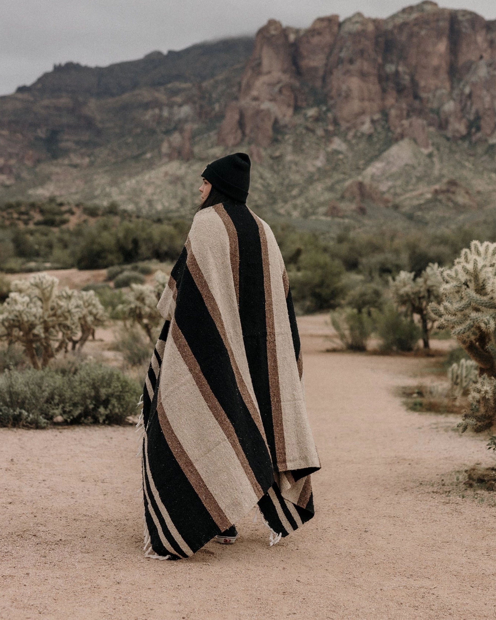 A person draped in the Faire TT Handwoven Blanket strolls along a desert path lined with cacti and shrubs, rocky mountains rising under a cloudy sky.