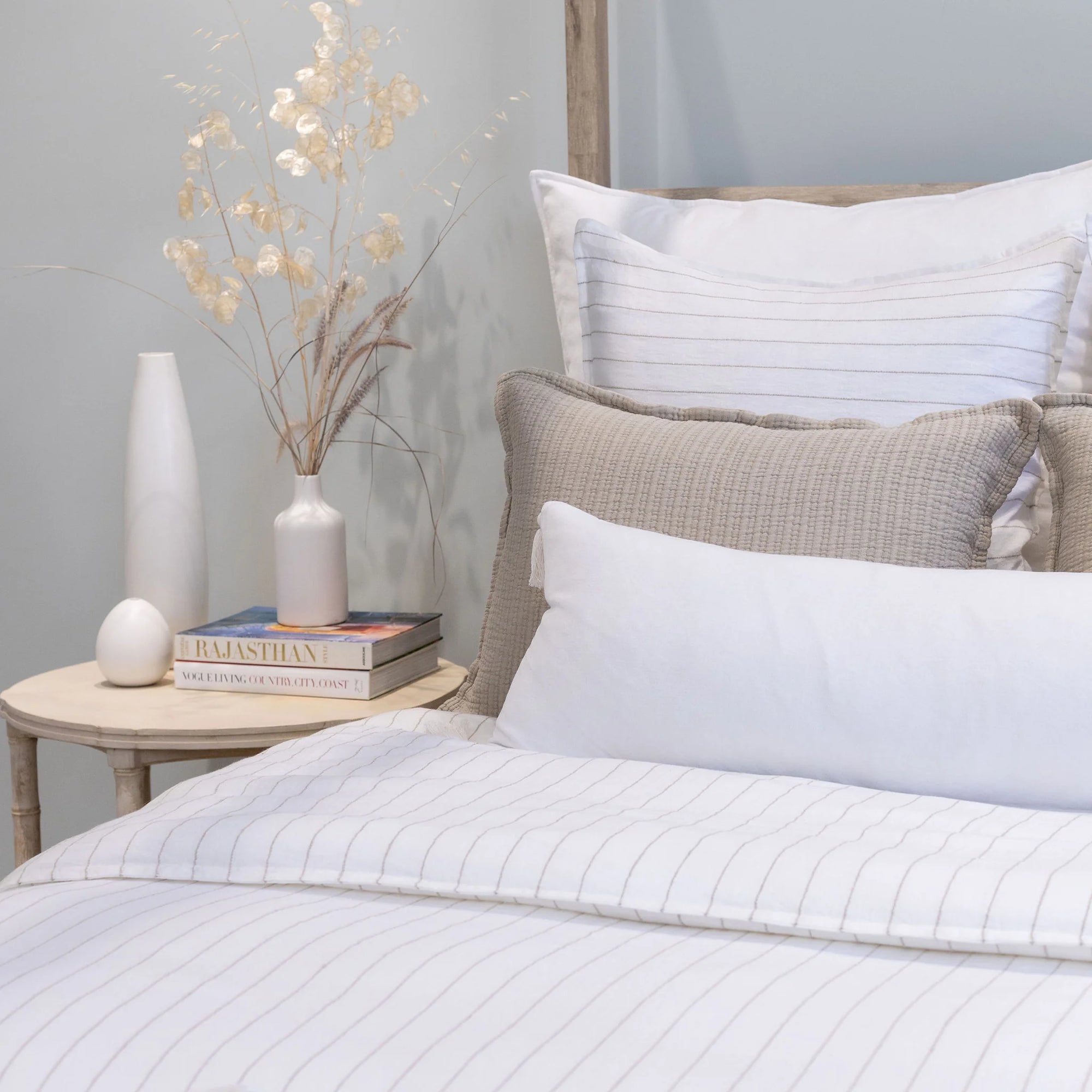 A cozy bedroom with a neatly made bed featuring sophisticated stripe bedding from the Lake Linen Collection and a Blake Big Pillow 28x36 by POM POM. Two stacked books and a white vase with dried flowers sit on a round wooden side table next to the bed, creating a setting with calm and minimalist decor.