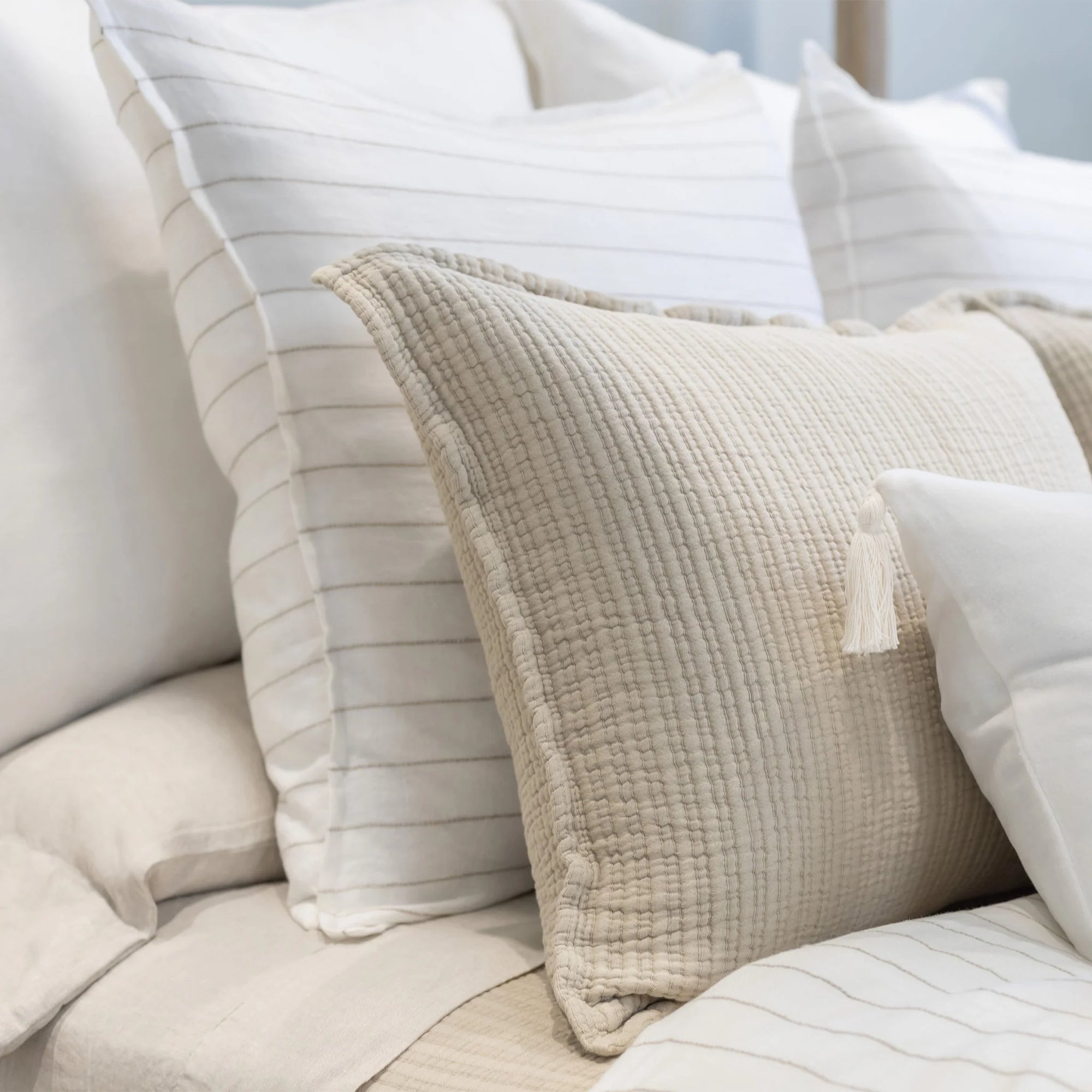 A neatly arranged bed adorned with a mix of textured pillows from POM POM&#39;s Lake Linen Collection, highlighted by the Blake Big Pillow 28x36 in beige with quilted tassels in the foreground. Smooth, white pillows featuring a sophisticated stripe pattern are positioned in the background, adding to the cozy and elegant atmosphere.