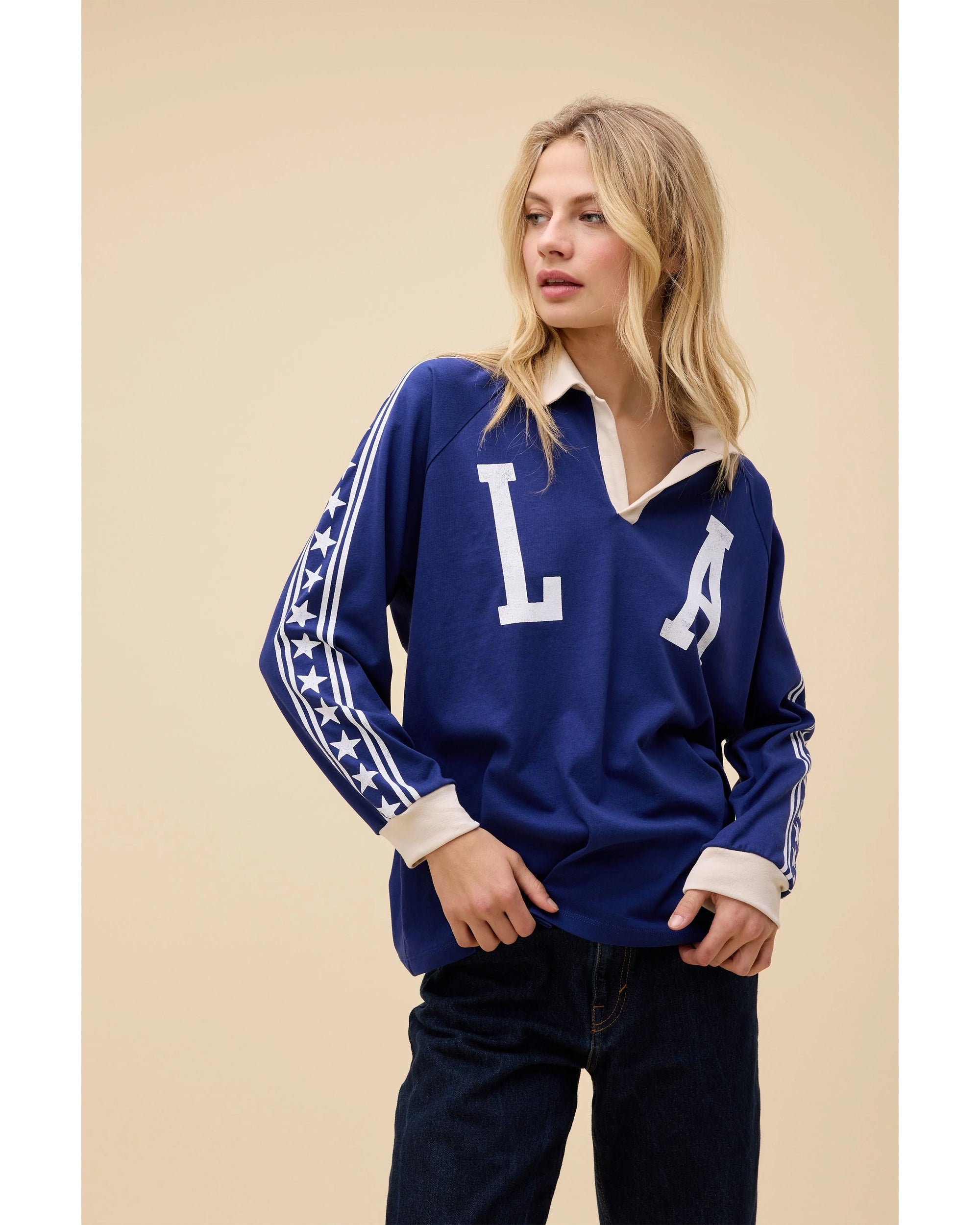 A blonde woman wears the DayDreamer Los Angeles Stars Rugby, an oversized blue raglan long sleeve with "LA" and white stars on the sleeves, paired with dark jeans against a beige background.