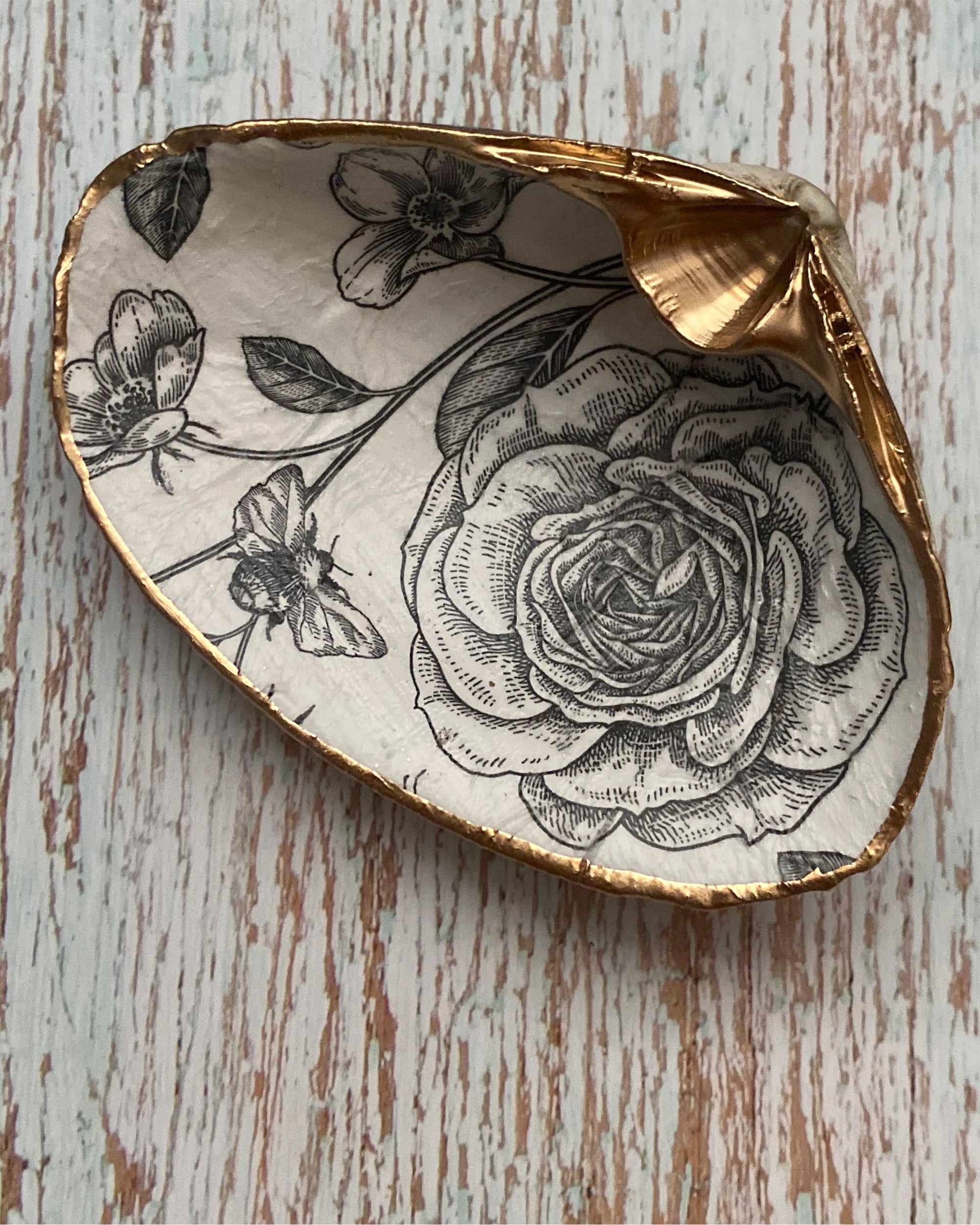 The Faire Black/White Roses Deco Shell features a paint-printed shell with gold-painted edges and a detailed black floral and butterfly design, ideal as a jewelry dish, shown on a light textured wooden background.