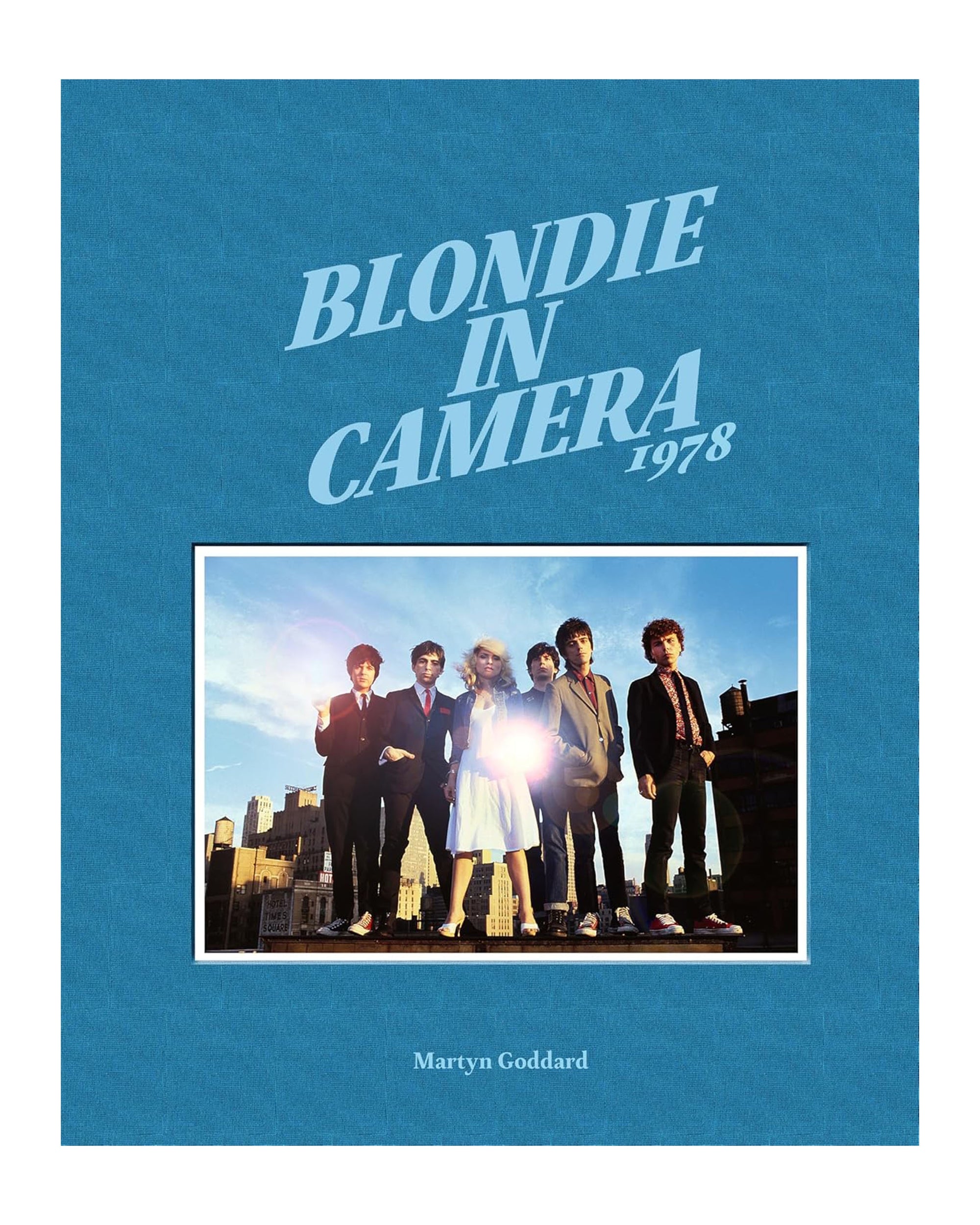 The "Blondie In Camera 1978" book cover from Simon & Schuster Inc. features a blue background, the title above a classic rooftop photo of Debbie Harry and Blondie, and Martyn Goddard’s name at the bottom.
