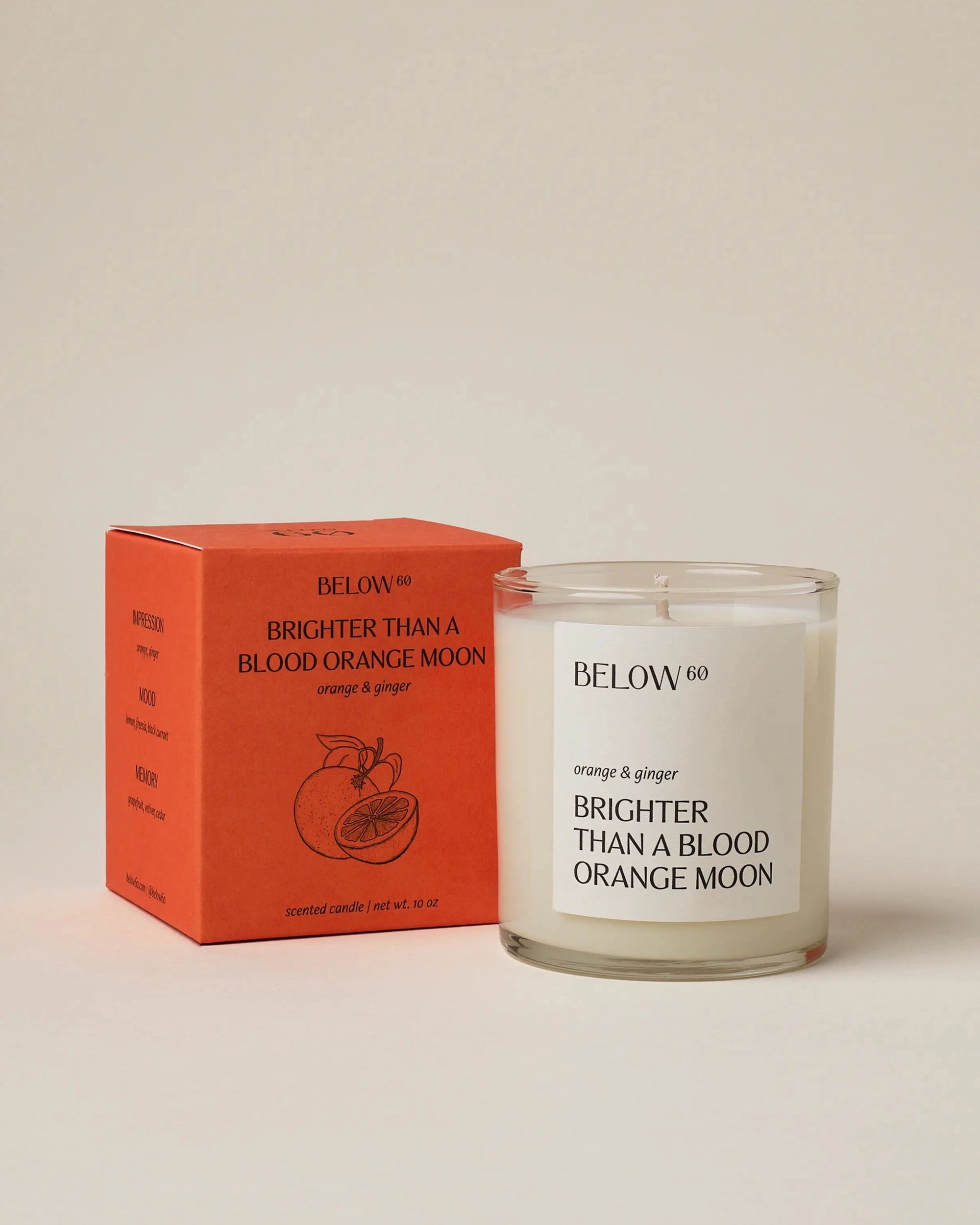 A glass "Below 60 Candle" by Faire stands beside its orange box, both labeled with “BRIGHTER THAN A BLOOD ORANGE MOON” and an orange illustration. This Leaping Bunny certified soy wax candle has a fresh orange & ginger scent.