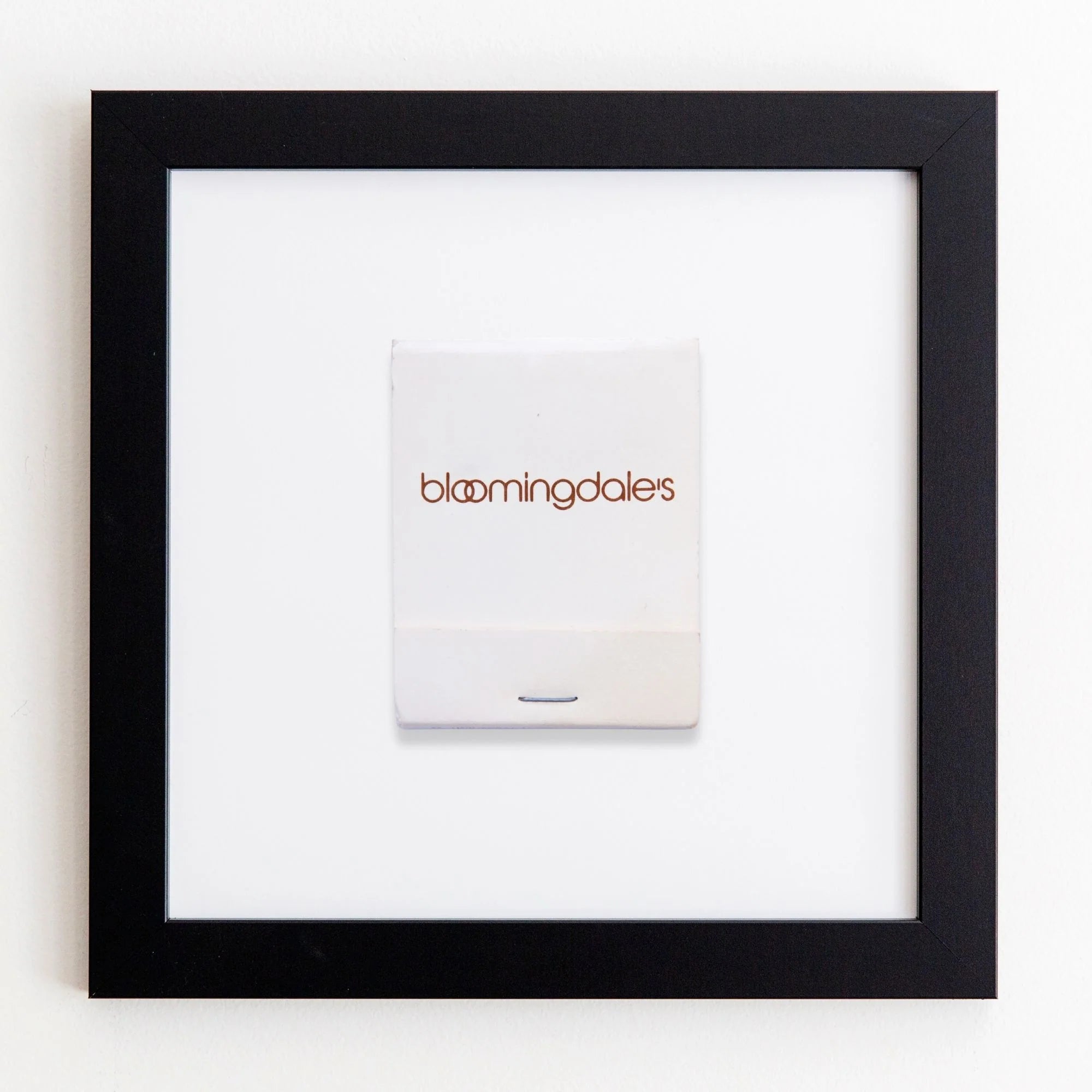 A framed Match South shopping bag, centered within a black acrylic frame, mounted on a white wall. The bag is plain white with the store&#39;s logo in lowercase black letters.