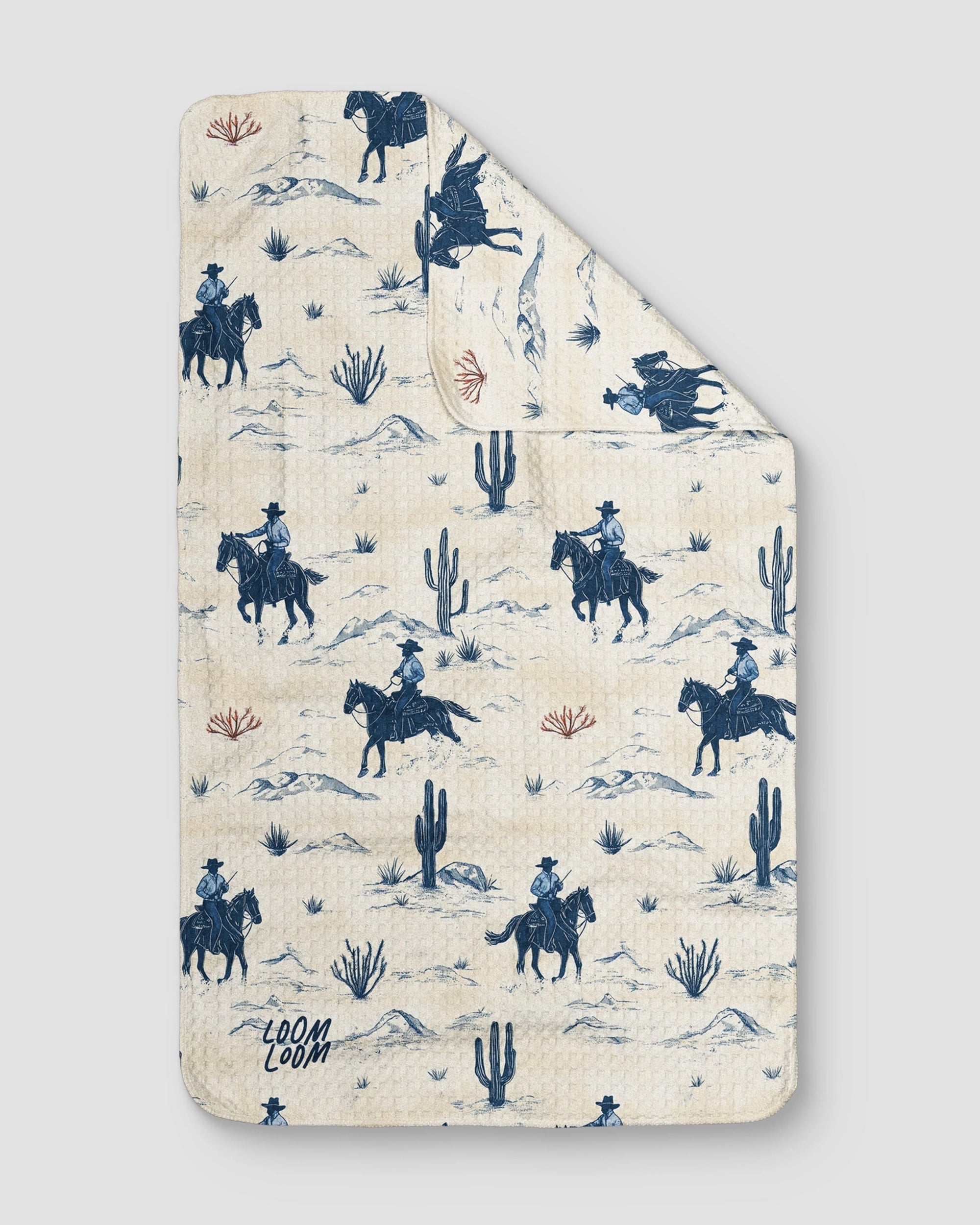 The “Cowboy Blues” quilt by Faire is cream-colored and features blue and beige cowboy and desert motifs, including cowboys on horseback, cacti, and shrubs. The brand name "Faire" appears in one corner.