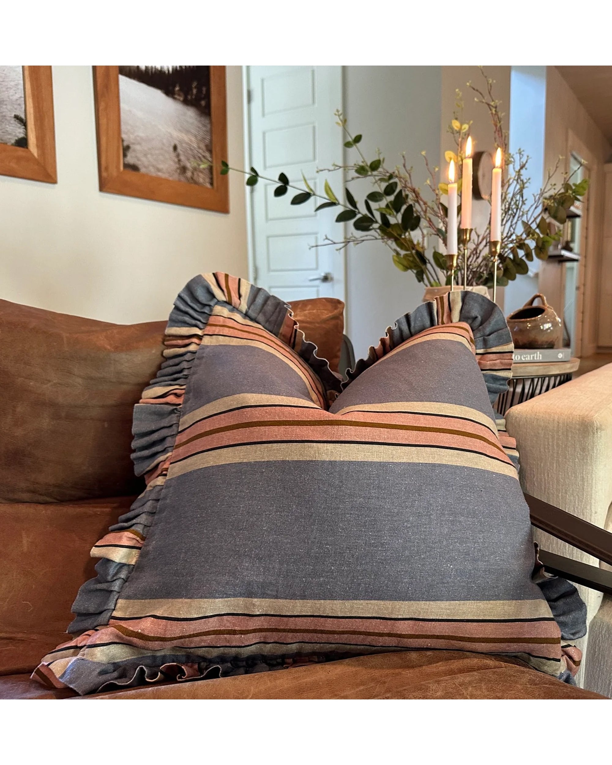 The Rennah Collective Foxy Blue Striped Ruffle Pillow 22x22 sits on a brown sofa, with candles, greenery, and framed wall art enhancing a cozy, bright living room accented by decorative throw pillows.