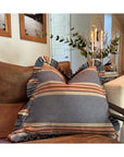 The Rennah Collective Foxy Blue Striped Ruffle Pillow 22x22 sits on a brown sofa, with candles, greenery, and framed wall art enhancing a cozy, bright living room accented by decorative throw pillows.