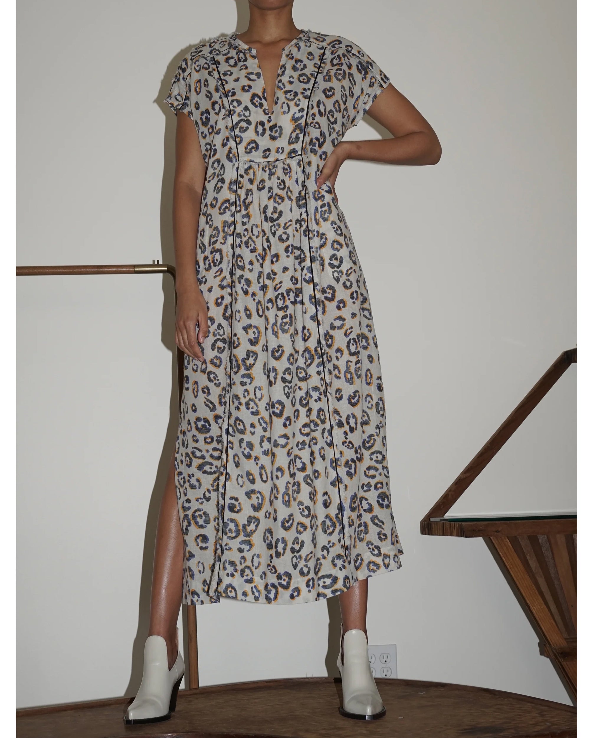 A person stands indoors wearing the Talitha Caftan by Raquel Allegra, a 100% cotton, short-sleeved, ankle-length blue and brown leopard print dress with a v-neckline and side slit, paired with white ankle boots.