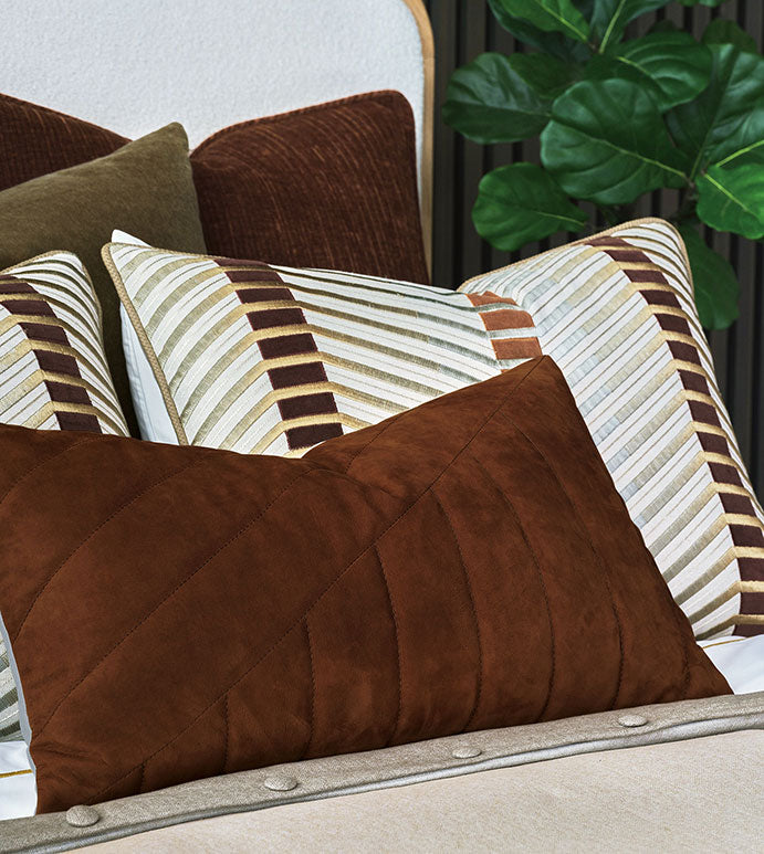 A bed with striped neutral pillows and a large Eastern Accents Rufus Nubuck Leather Pillow in front, set against a channeled headboard with a green leafy plant in the background.