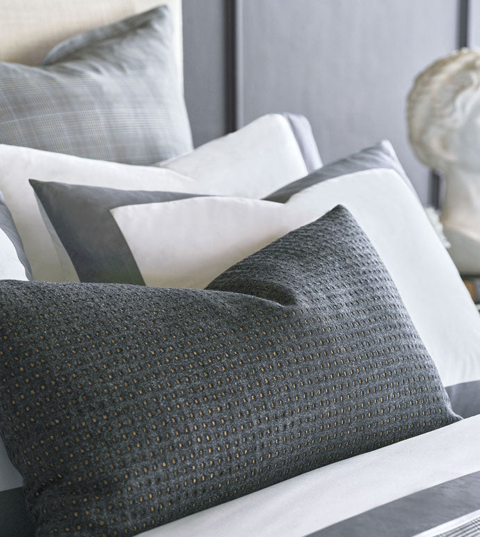 A neatly arranged bed features layered pillows in shades of gray and white, including the striking OCTAVIUS WOOL pillow by Eastern Accents, all set against a dark headboard with a blurred decorative sculpture in the background.