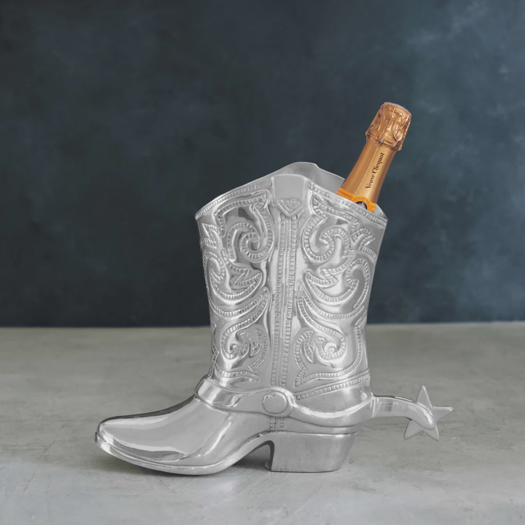 A silver, ornate Beatriz Ball Cowboy Boot Wine Bucket crafted from aluminum alloy holds a bottle of champagne, with the top of the bottle visible. The scene is set against a dark, textured background on a light-colored surface, evoking a sense of Western richness.