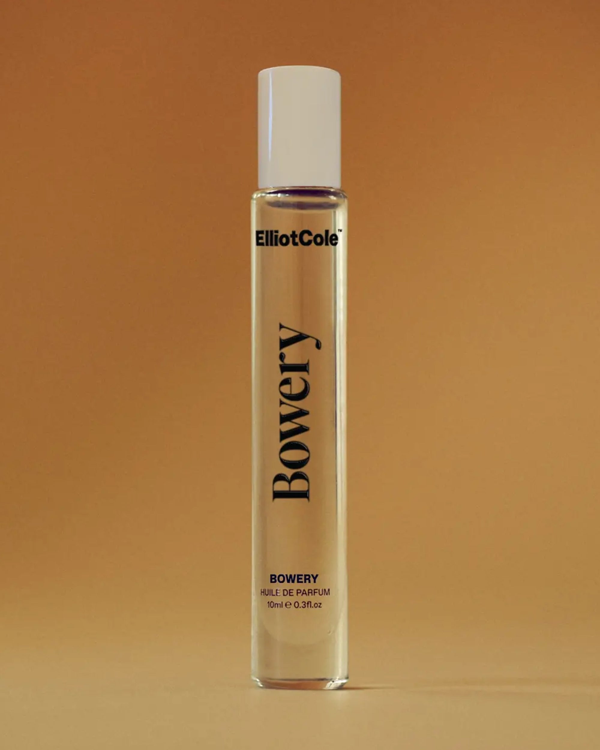 A clear glass 10 ml Bowery Roll-On perfume bottle stands upright against a warm brown background, featuring a white cap and light liquid. This unisex scent captures the spirit of New York City in an elegant, modern design.