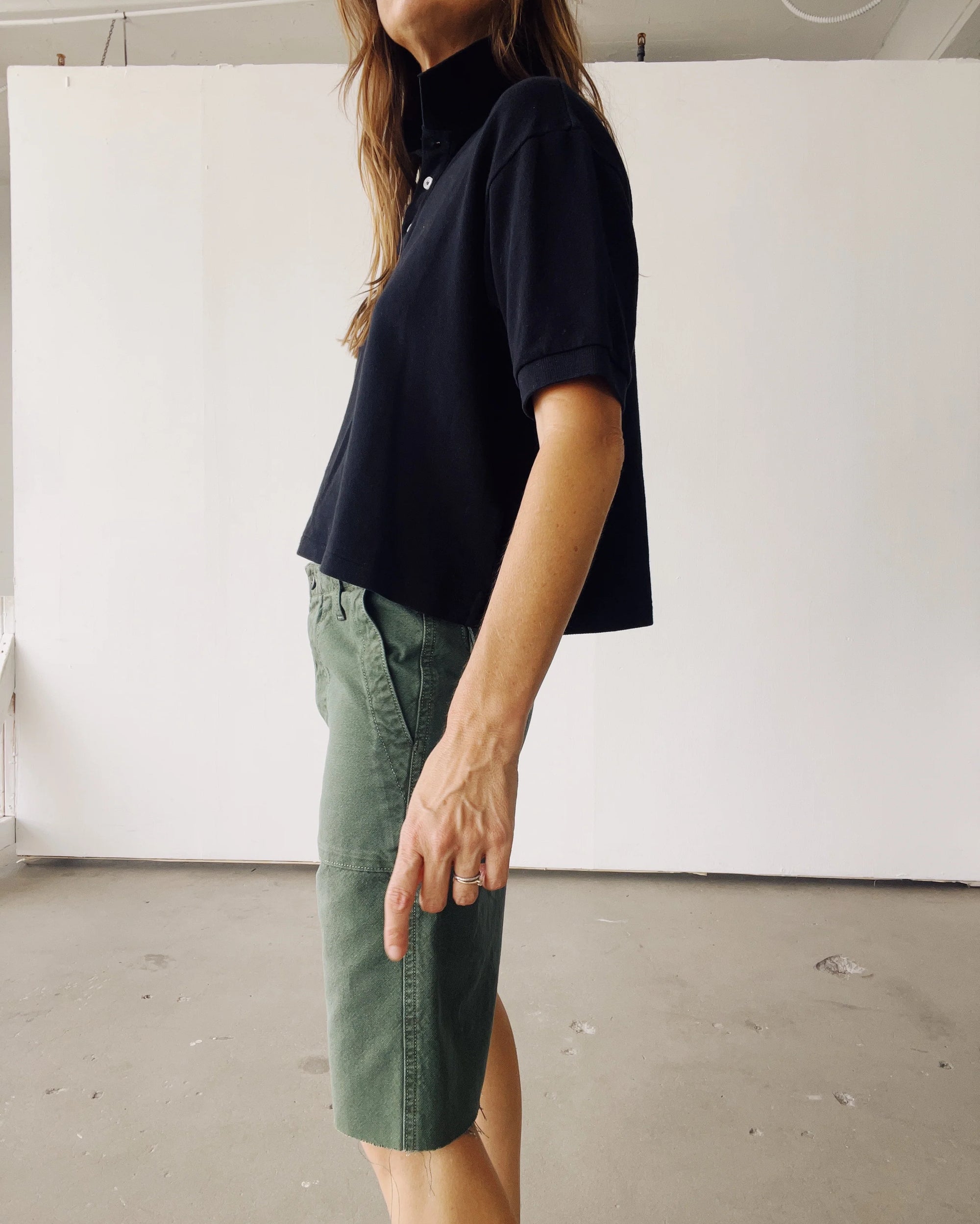 Indoors, a person stands in profile wearing Hey Gang’s The Boxy Polo in black with green knee-length shorts. Their arm is relaxed by their side, set against plain white walls and a concrete floor.