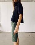 Indoors, a person stands in profile wearing Hey Gang’s The Boxy Polo in black with green knee-length shorts. Their arm is relaxed by their side, set against plain white walls and a concrete floor.