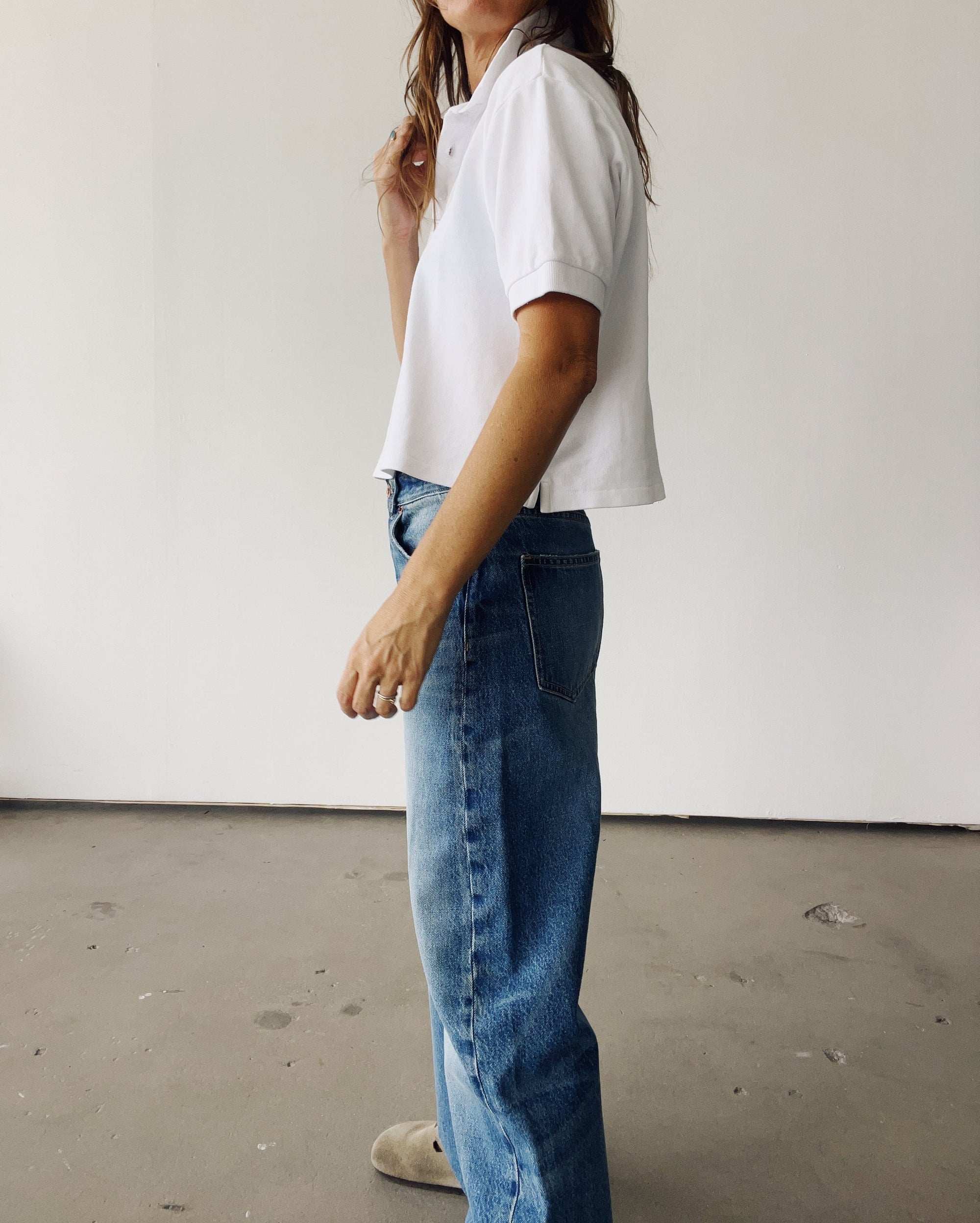 A person stands indoors against a white wall, wearing Hey Gang's The Boxy Polo in short sleeves and high-waisted blue jeans. Only part of their face is visible, with loose brown hair.
