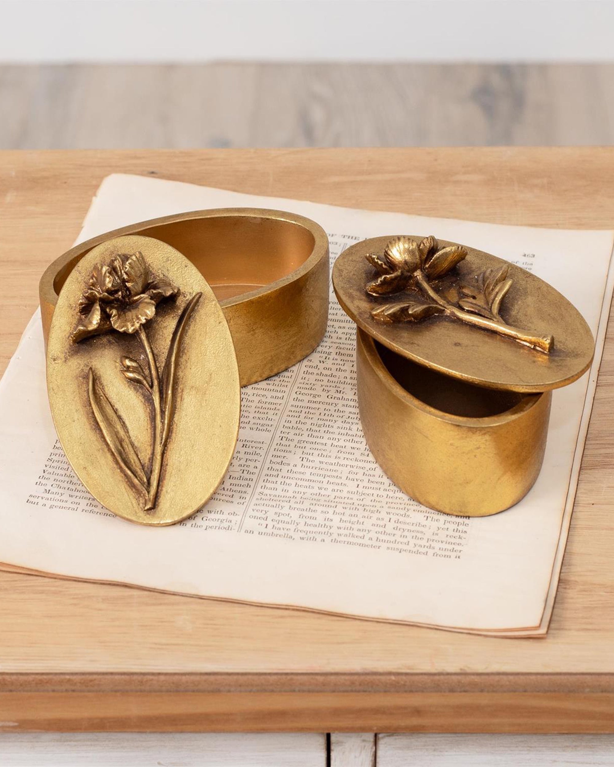 Two small Faire Brass Tone Floral Boxes with embossed floral lids are displayed on an open book and wood surface; one keepsake box is open, showing its empty interior. Vintage-inspired design adds charm to any décor.
