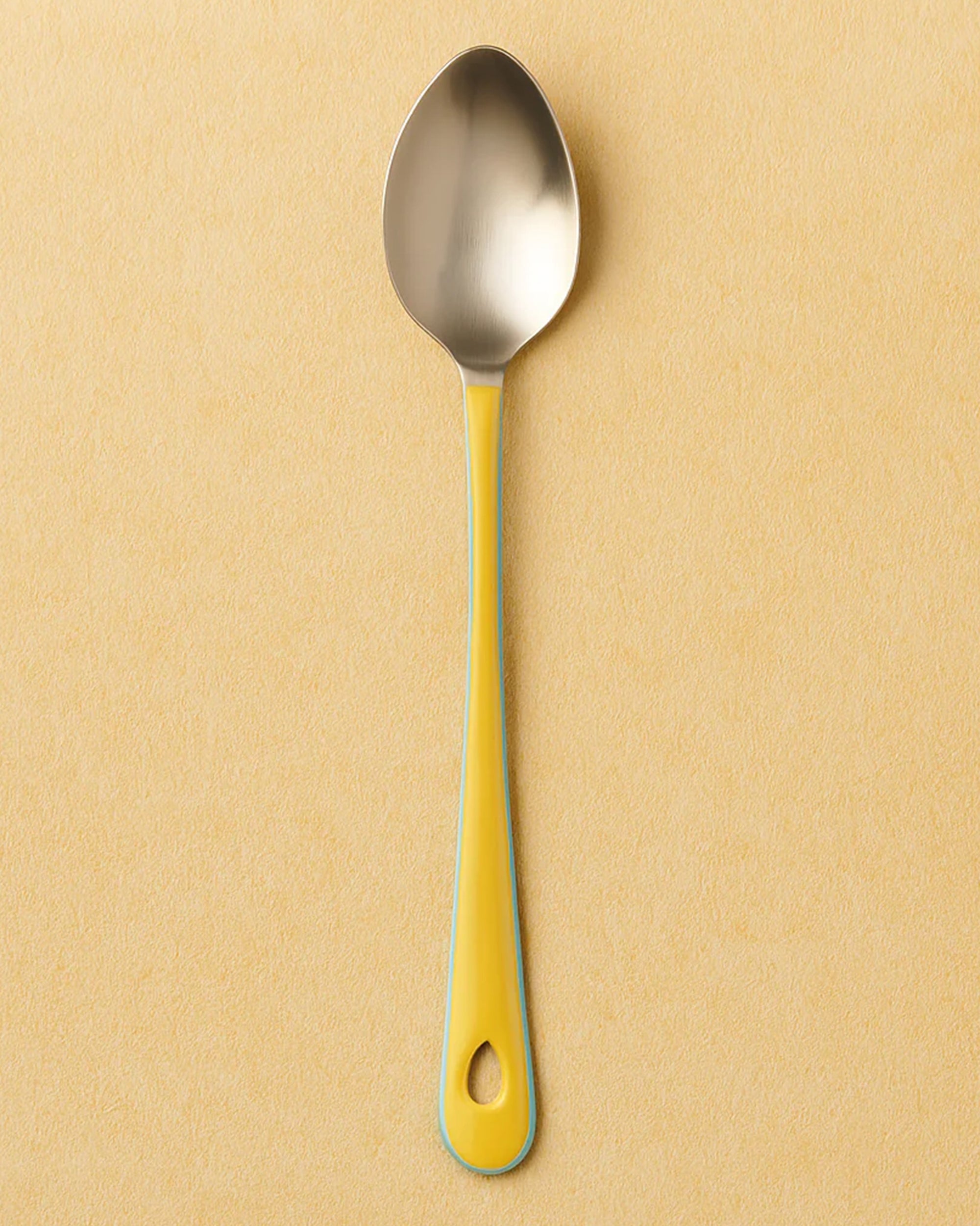 The Harlow Bright Mixing Spoon by Be Home is a stainless steel serveware piece with a yellow handle and vintage style, featuring a small end hole and shown on a light beige surface.