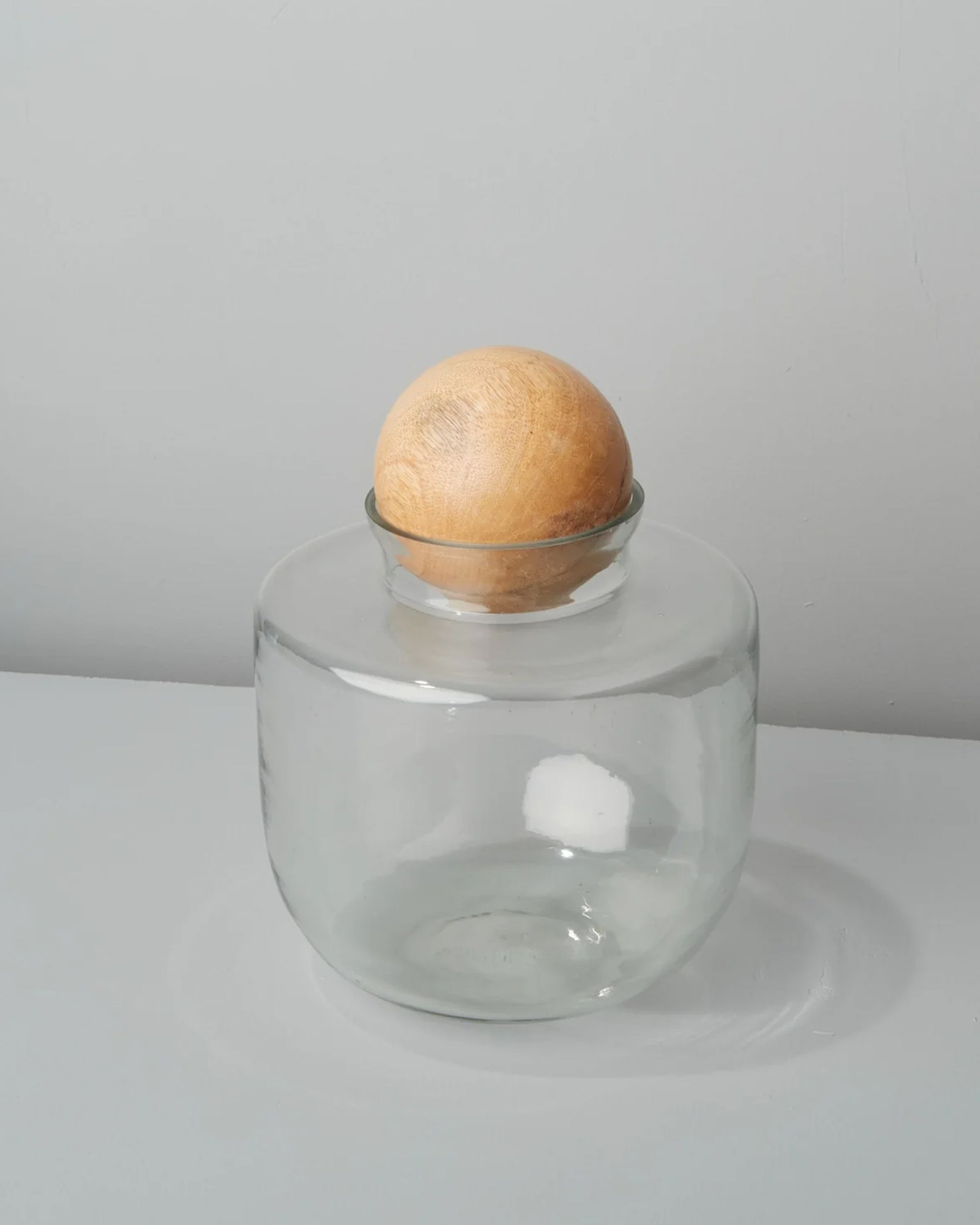 The Brisbane Short Canister by Be Home features clear recycled glass and a round, ethically sourced mango wood lid, bringing charm and sustainability to your home decor. Displayed on a gray surface with a light gray background.