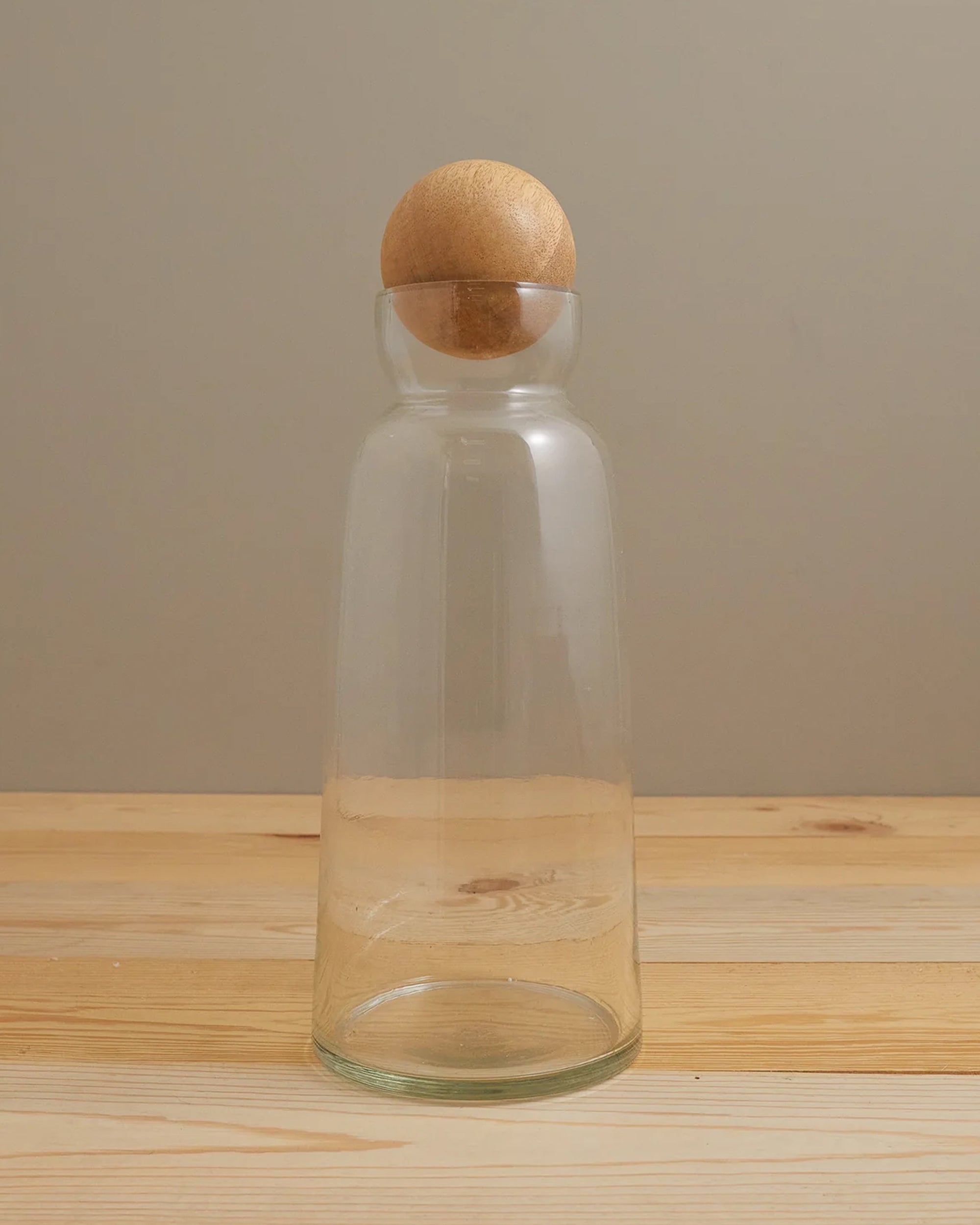 The Be Home Brisbane Decanter Tall, crafted from clear recycled glass and topped with a round mango wood stopper, rests on a light wooden surface against a plain beige backdrop.