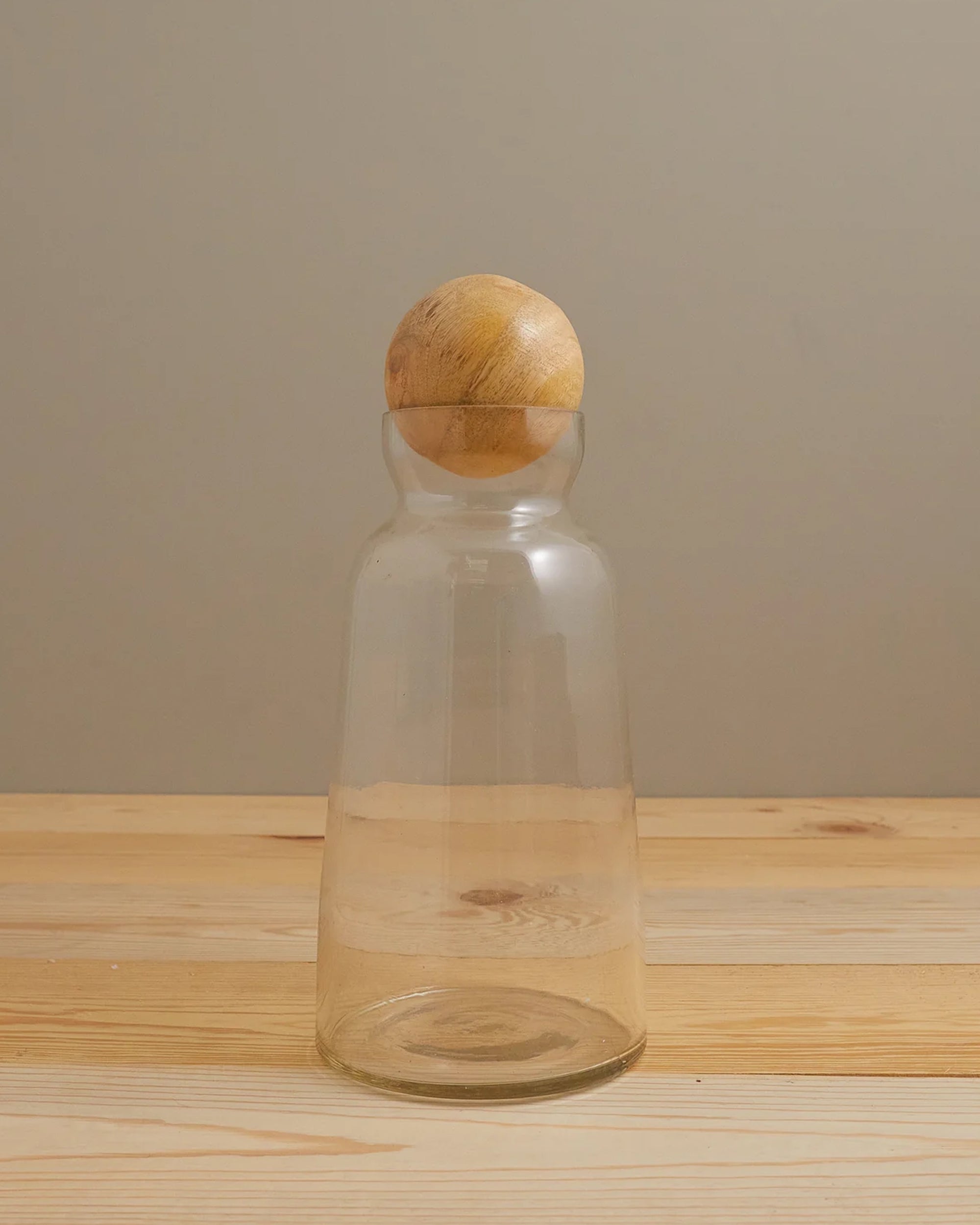 The Be Home Brisbane Decanter M features clear recycled glass with a wide base and narrow neck, topped by a round mango wood lid, and is shown on a light wood surface against a beige background.