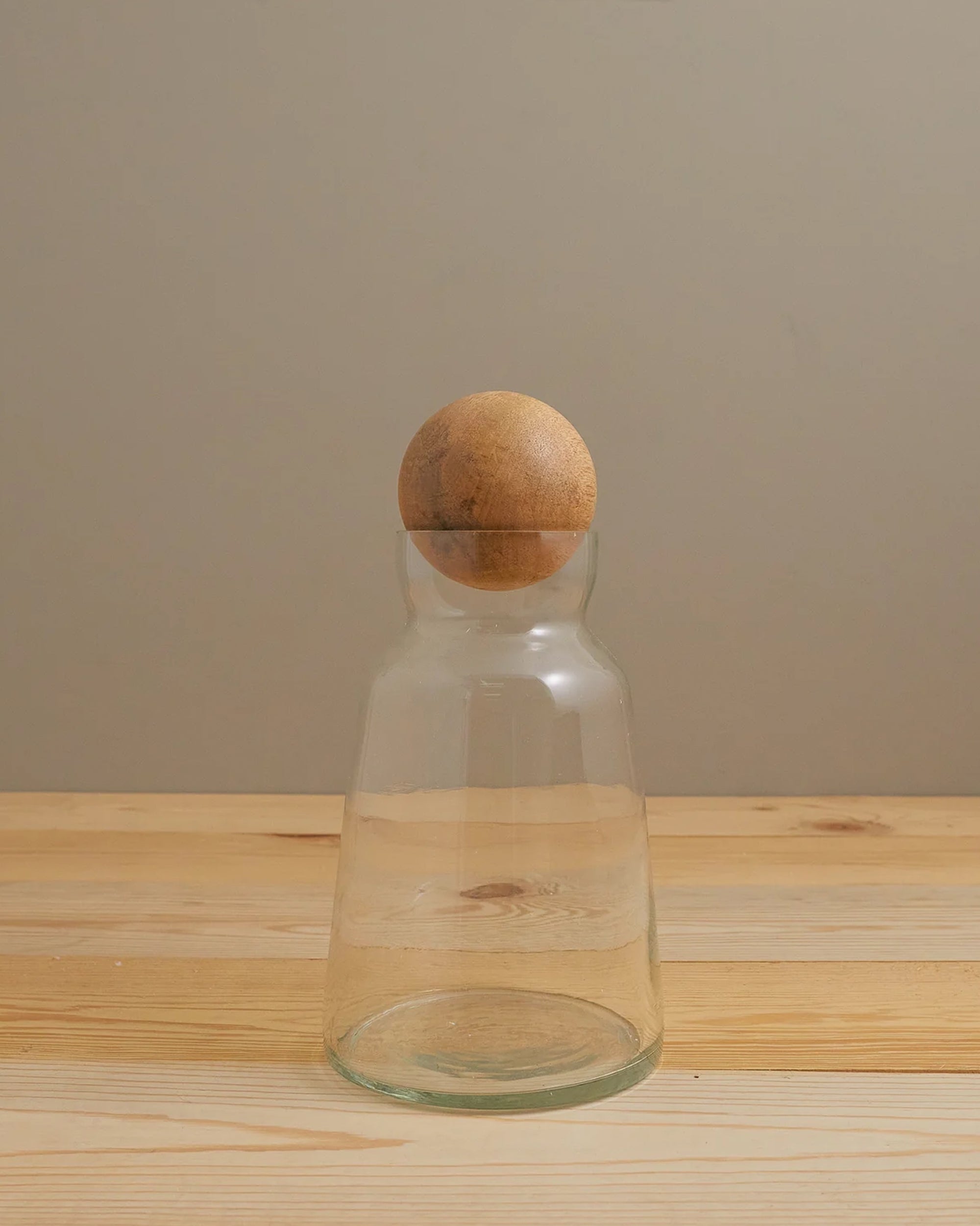 The Brisbane Decanter Short by Be Home features clear recycled glass and a round mango wood stopper, providing a fresh twist on classic decanters. Shown on a light wooden surface with a plain beige backdrop.