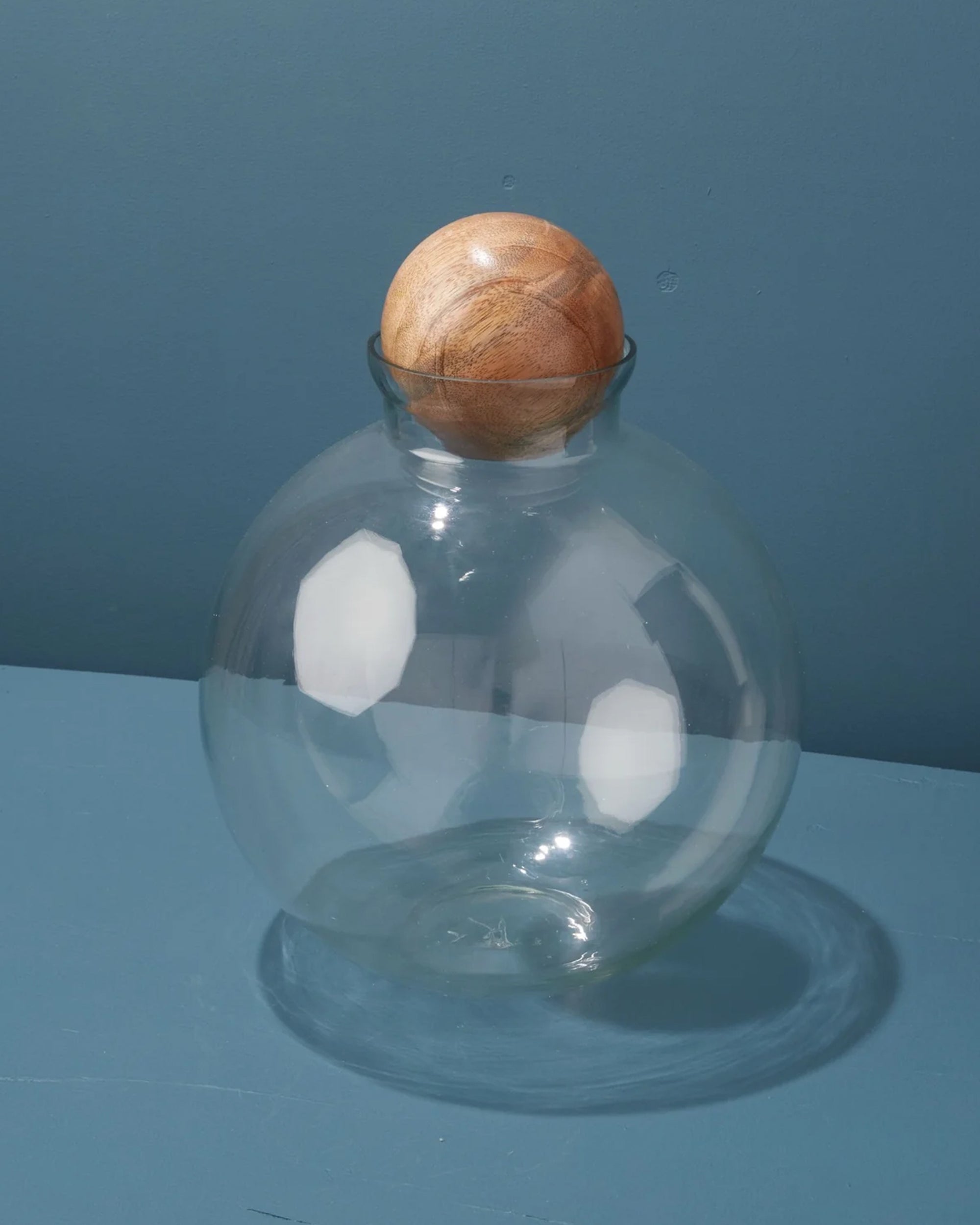 The Be Home Brisbane Spherical Canister, featuring a clear recycled glass jar and a round mango wood lid, rests on a blue surface against a blue wall. Light reflects off the glass, highlighting its eco-friendly kitchen style.
