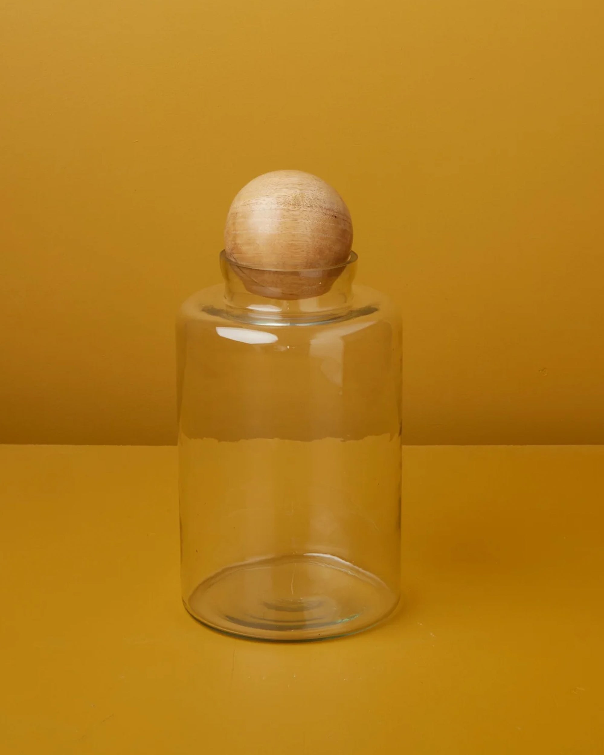 The Be Home Brisbane Tall Canister, featuring clear recycled glass and a round mango wood lid, sits on a mustard yellow surface with a matching mustard yellow background.