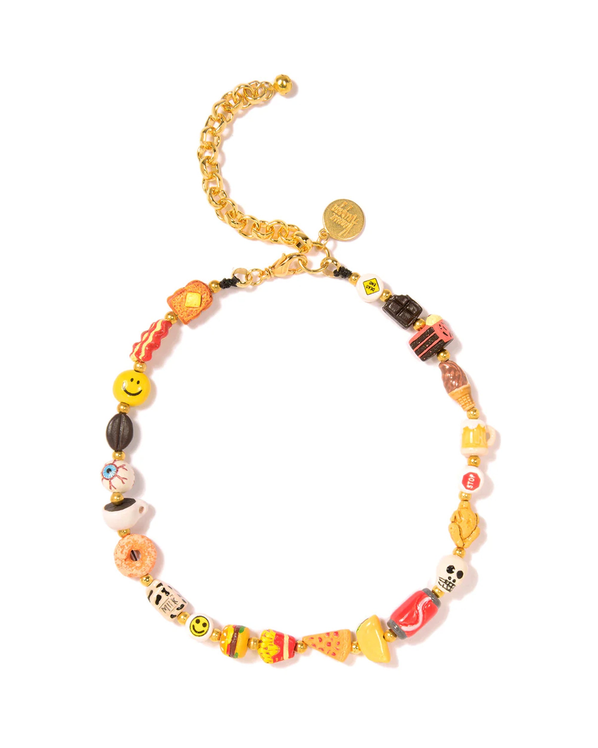 The Breakfast Lunch Dinner Necklace 14" by Venessa Arizaga features colorful, mismatched ceramic charms shaped like foods, smiley faces, flowers, and more on a gold chain with a small round gold tag near the clasp.