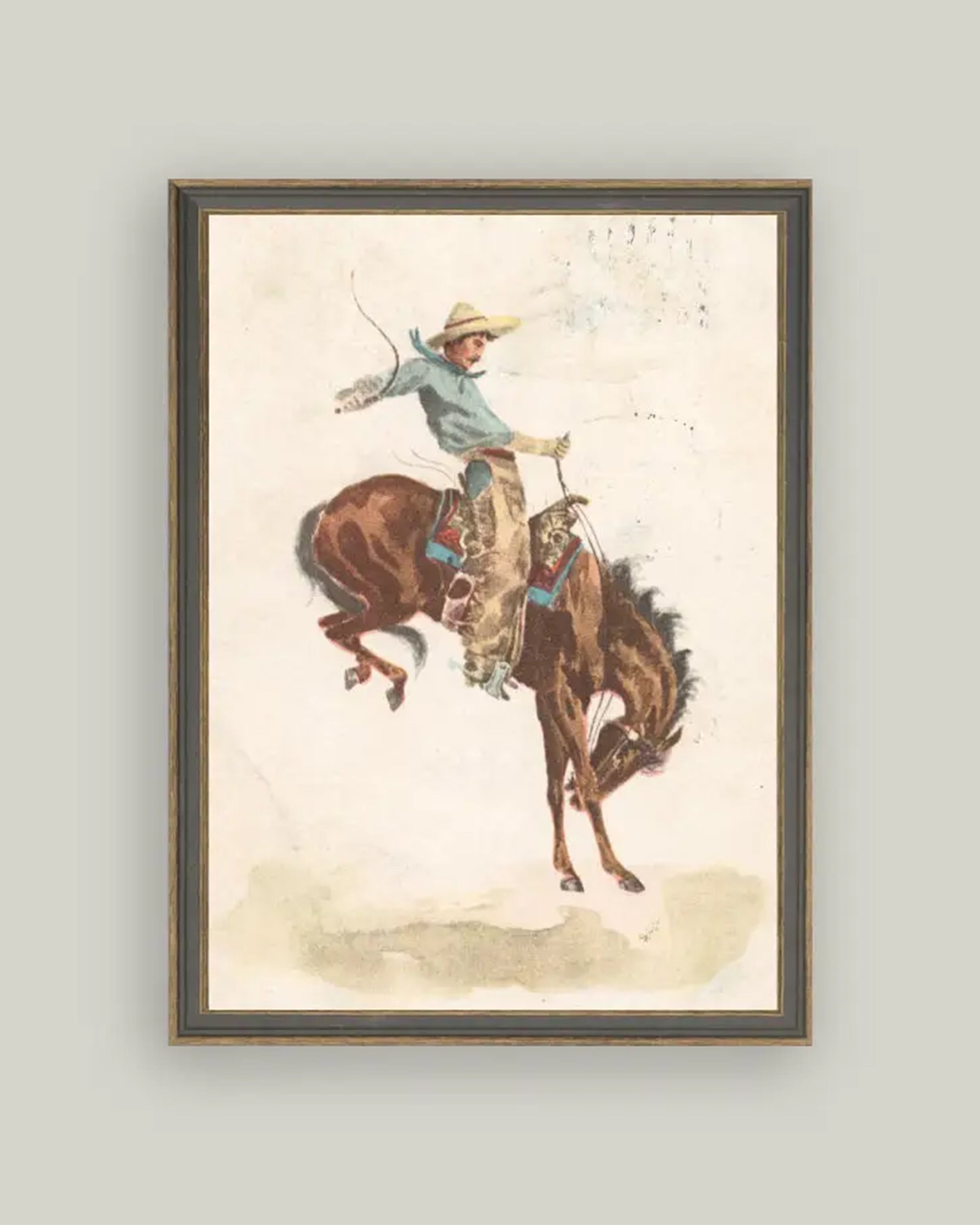 The Bucking Bronco Art by Petal Lane Home features a cowboy in a hat, blue shirt, and tan chaps riding a brown horse with a decorated saddle on a neutral background, set in an ornate handcrafted frame. Proudly made in the USA.