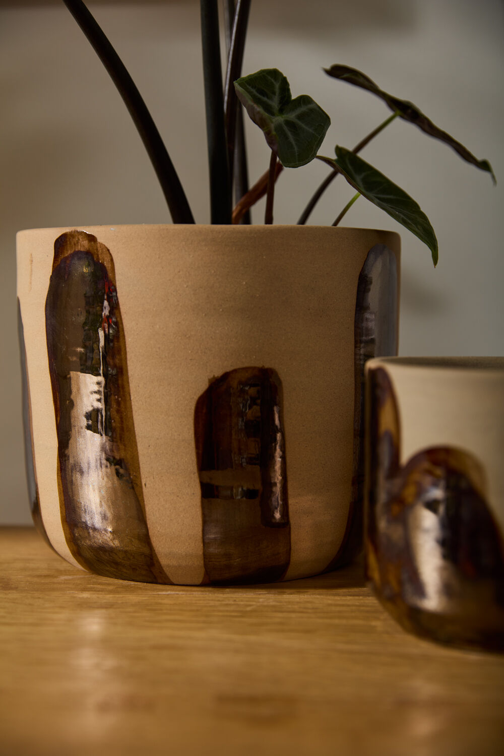 The Bronze Brushstroke Pot by Accent Decor is a handcrafted beige ceramic piece with abstract brown glossy designs, holding a leafy plant. It sits on a wooden surface next to a matching smaller item for decorative appeal.