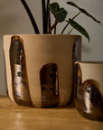 The Bronze Brushstroke Pot by Accent Decor is a handcrafted beige ceramic piece with abstract brown glossy designs, holding a leafy plant. It sits on a wooden surface next to a matching smaller item for decorative appeal.