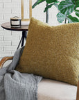 A cozy living room features the Eastern Accents Marl Mustard Pillow with a textured finish and invisible zipper, paired with a soft gray blanket, a round side table holding a candle, and a leafy green plant against a white brick wall.