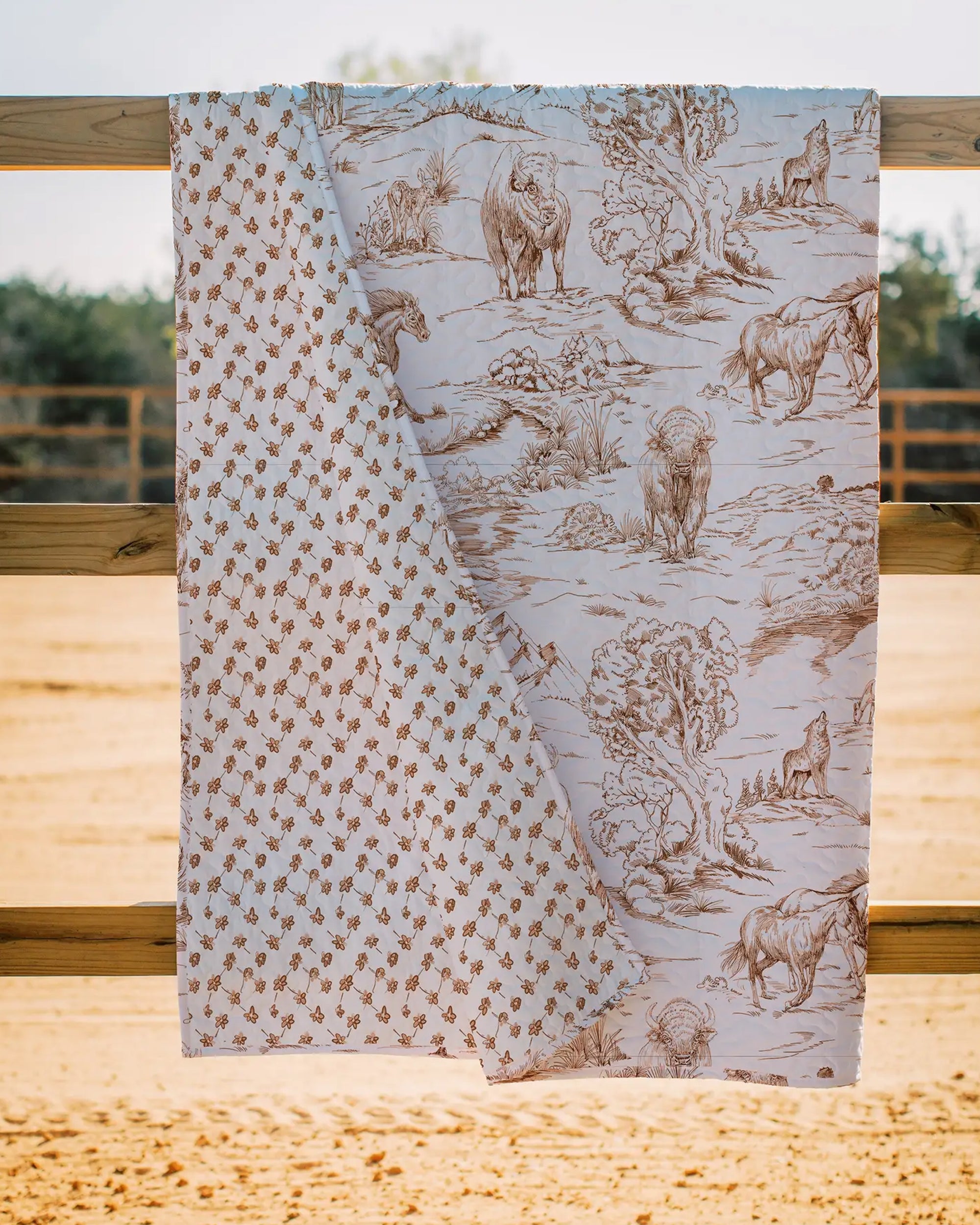 The Faire Buffalo Toile Brown Quilt is draped over a wooden fence, displaying brown wildlife and nature scenes on one side and a small repeating floral or animal print on the other. Set outdoors with sand and trees, it's ideal for cozy bedding.