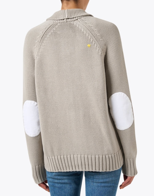 A person is wearing a beige knit Joy Sweater Coat by Kerri Rosenthal, which has white elbow patches, paired with blue jeans. This sweater features a yellow logo near the neckline on the back, set against a plain white background.