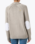 A person is wearing a beige knit Joy Sweater Coat by Kerri Rosenthal, which has white elbow patches, paired with blue jeans. This sweater features a yellow logo near the neckline on the back, set against a plain white background.