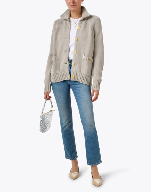 A person wearing the beige "Joy Sweater Coat" by Kerri Rosenthal with multicolor embroidery, paired with a white shirt, blue jeans, and beige shoes, holds a small silver handbag. They are standing in front of a white background.