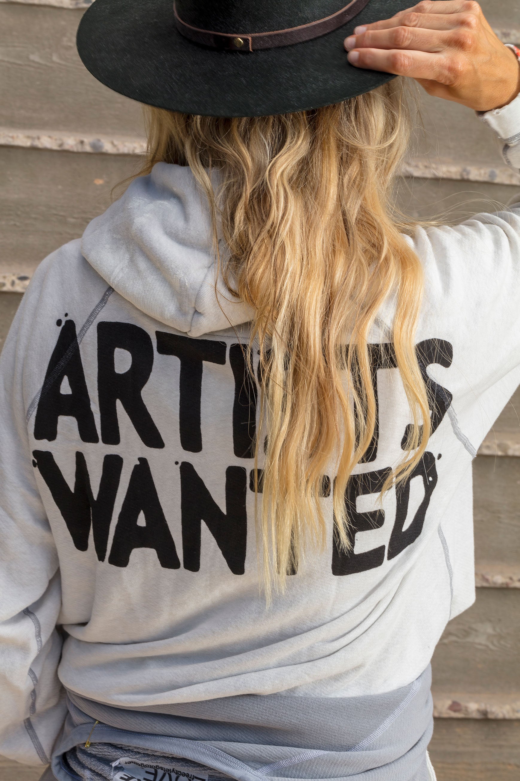 A woman from behind wearing a gray hoodie with the text "ARTISTS WANTED" in bold lettering, touching the brim of her black hat, with wavy blonde hair visible.