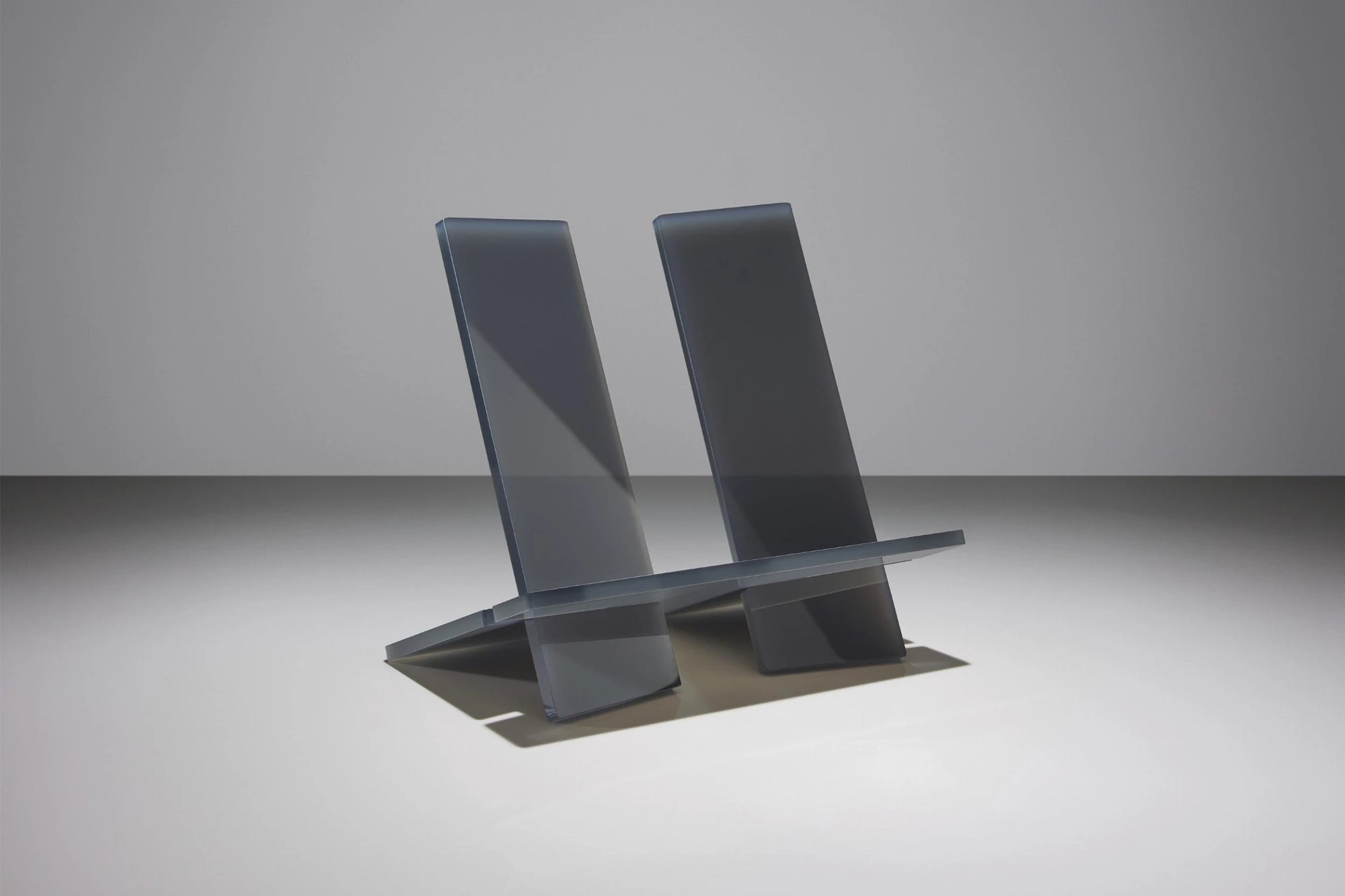 The Ingram Book Company’s Bookstand Md is a minimalist, modern chair crafted from translucent gray material with two angled backrests and a flat seat, set against a plain, light gray background.