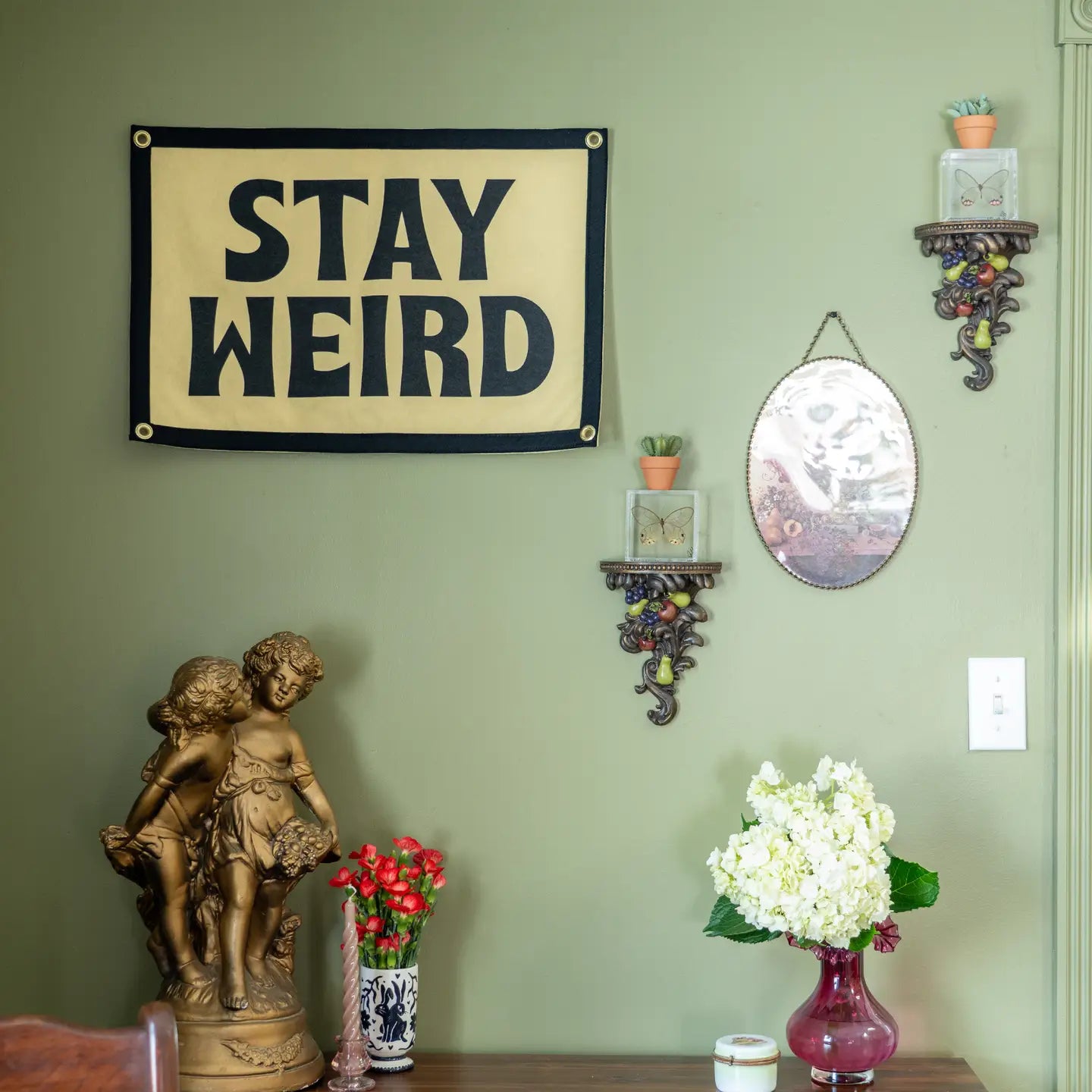 A green wall with bold energy displays an Oxford Pennant "Stay Weird Camp Flag" (16x24), an oval mirror, ornate shelves with candles, a statue of two children, and vases of red and white flowers on a wooden table.