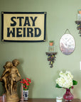 A green wall with bold energy displays an Oxford Pennant "Stay Weird Camp Flag" (16x24), an oval mirror, ornate shelves with candles, a statue of two children, and vases of red and white flowers on a wooden table.