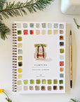 The Faire Watercolor Workbook is a spiral-bound notebook with watercolor squares and a painted camper on the cover. Beginner-friendly, it’s shown with fir branches, yellow flowers, and a paintbrush on a white surface.