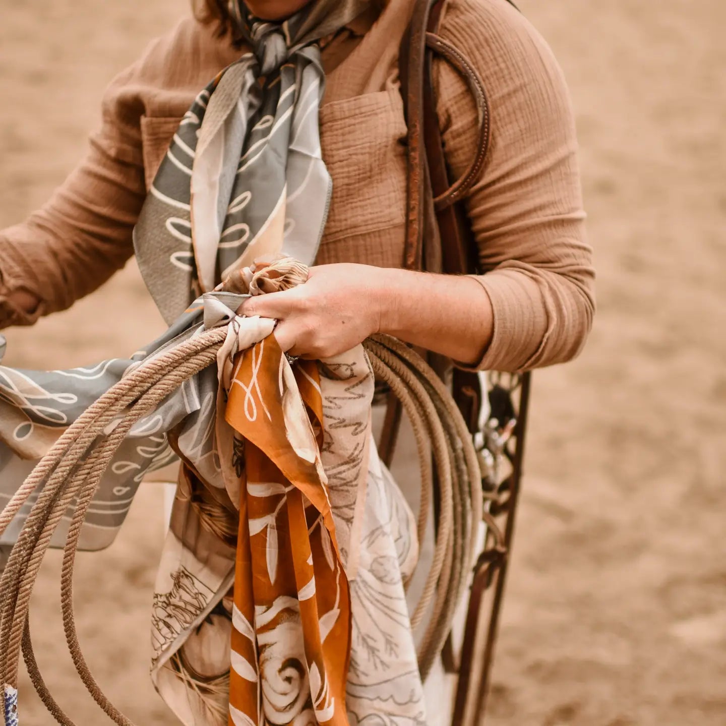 A person in a brown shirt with a patterned cowgirl accessory holds several ropes and colorful Wild Rag Silk Scarves by Faire, standing on sandy ground. Their face is not visible.