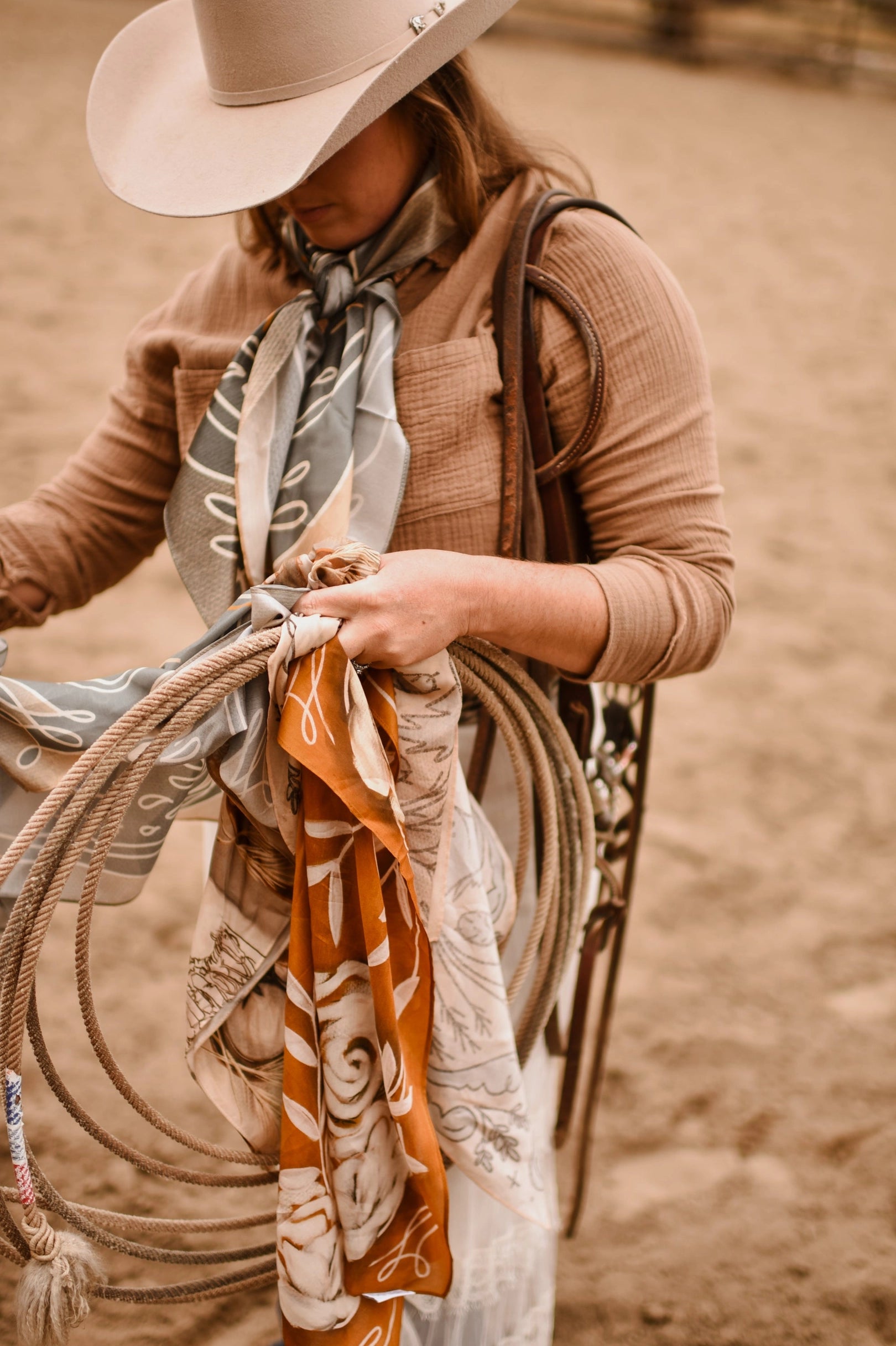 Wearing a wide-brimmed hat and the Faire Rustic Wild Rose Western Silk Scarf, a person stands on sandy ground holding several ropes and colorful bandanas, their face partially hidden by the hat.
