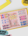 A vibrant yellow box of Faire's "Days of The Week Press on Nail Kit" features colorful nails with designs like rainbows, smiley faces, and leopard prints. The box boasts "7 Super Styles" and "Pre-Applied Adhesive.