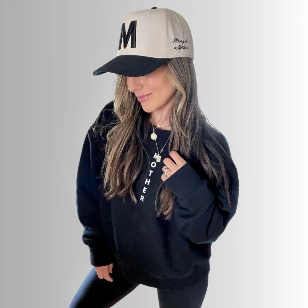 A woman wears Faire's The M Hat, a black embroidered cap with a bold "M", paired with a black sweatshirt, leggings, and a "MOTHER" necklace. Her wavy hair completes this strong look against a light gradient background.