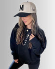 A woman wears Faire's The M Hat, a black embroidered cap with a bold "M", paired with a black sweatshirt, leggings, and a "MOTHER" necklace. Her wavy hair completes this strong look against a light gradient background.